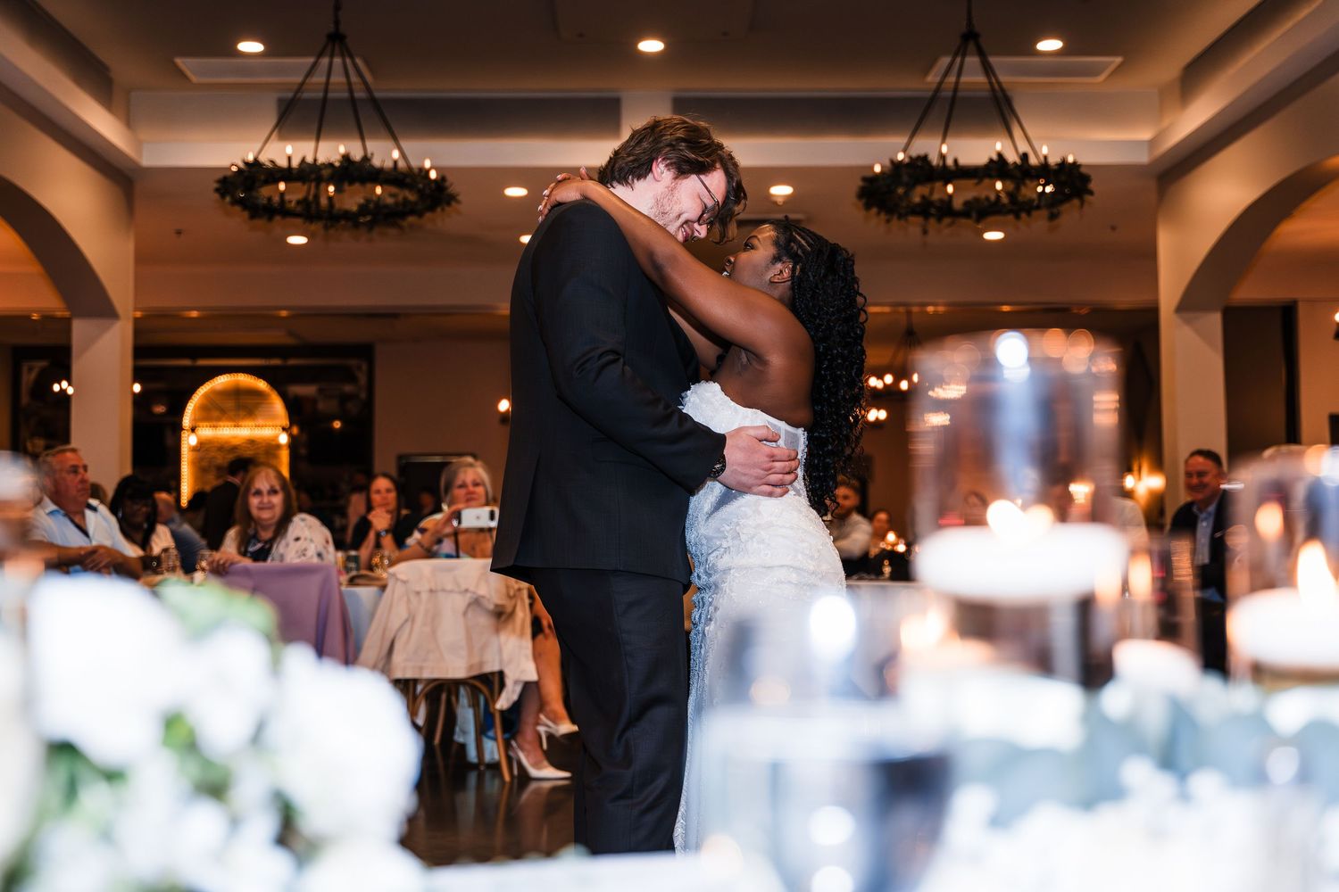 Bride and groom embracing during first dance in a beautifully lit wedding reception ballroom.