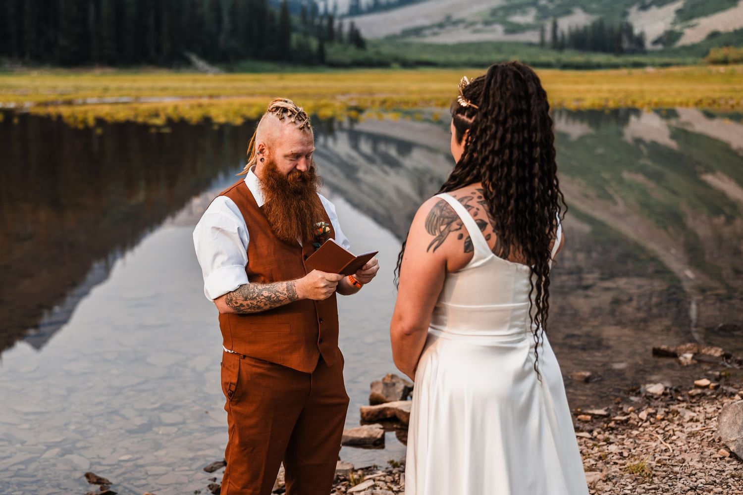 Groom in brown vest reads wedding vows to bride in white dress during outdoor elopement by a lake.