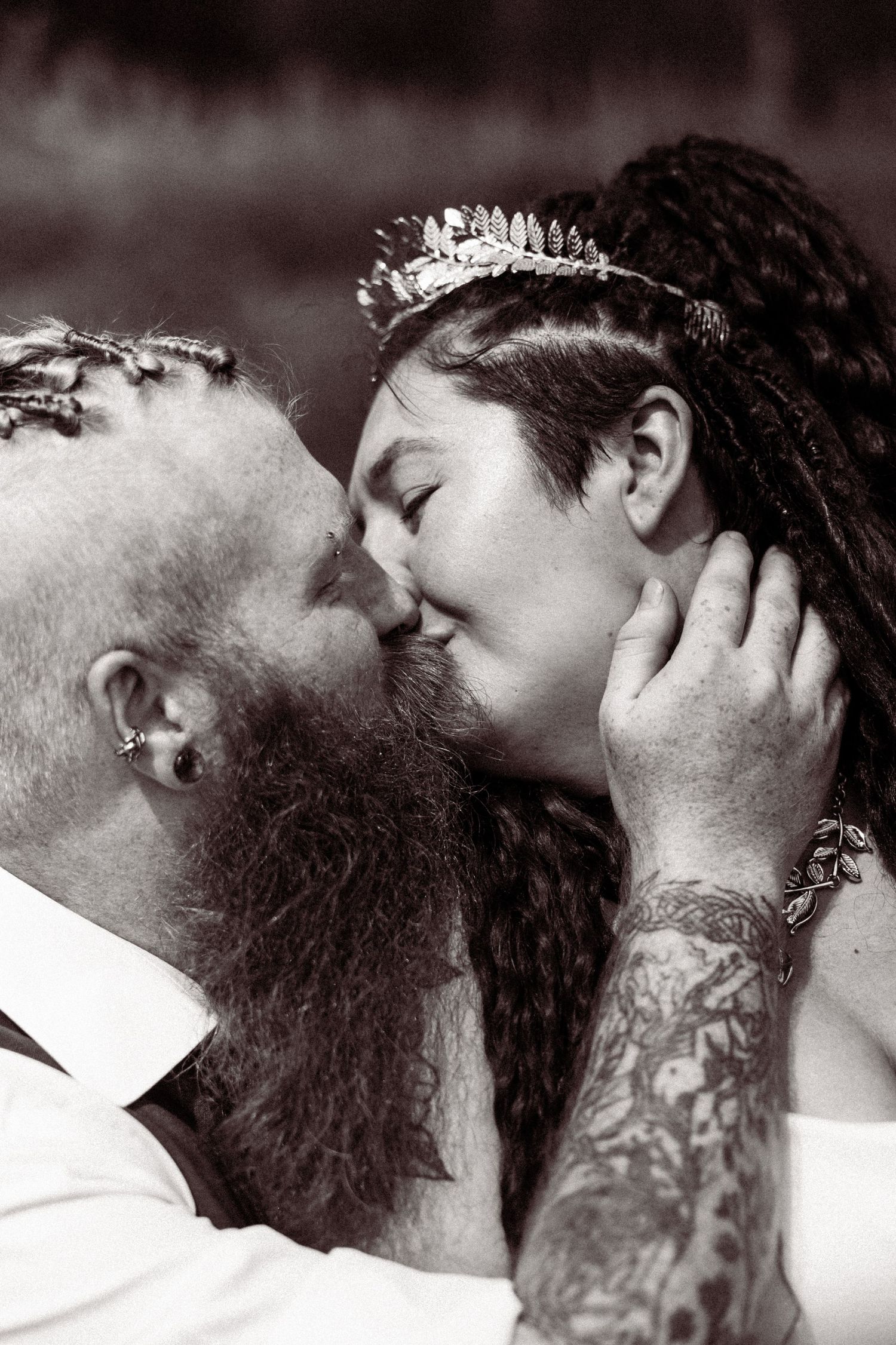 Black and white photo of tattooed groom with long beard kissing bride wearing a leaf crown tiara.
