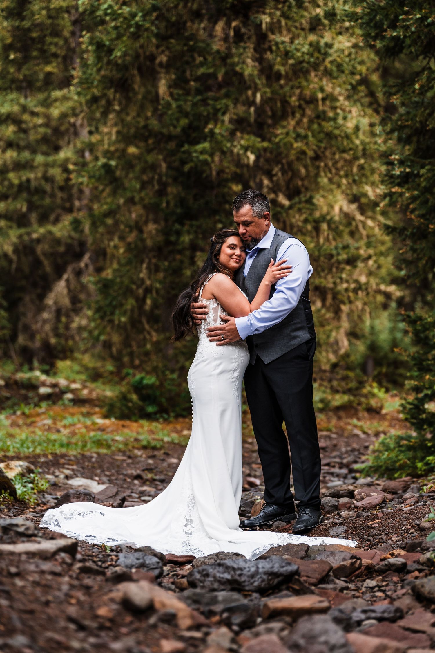 Bride and groom share a tender embrace on rocky ground surrounded by dense forest.