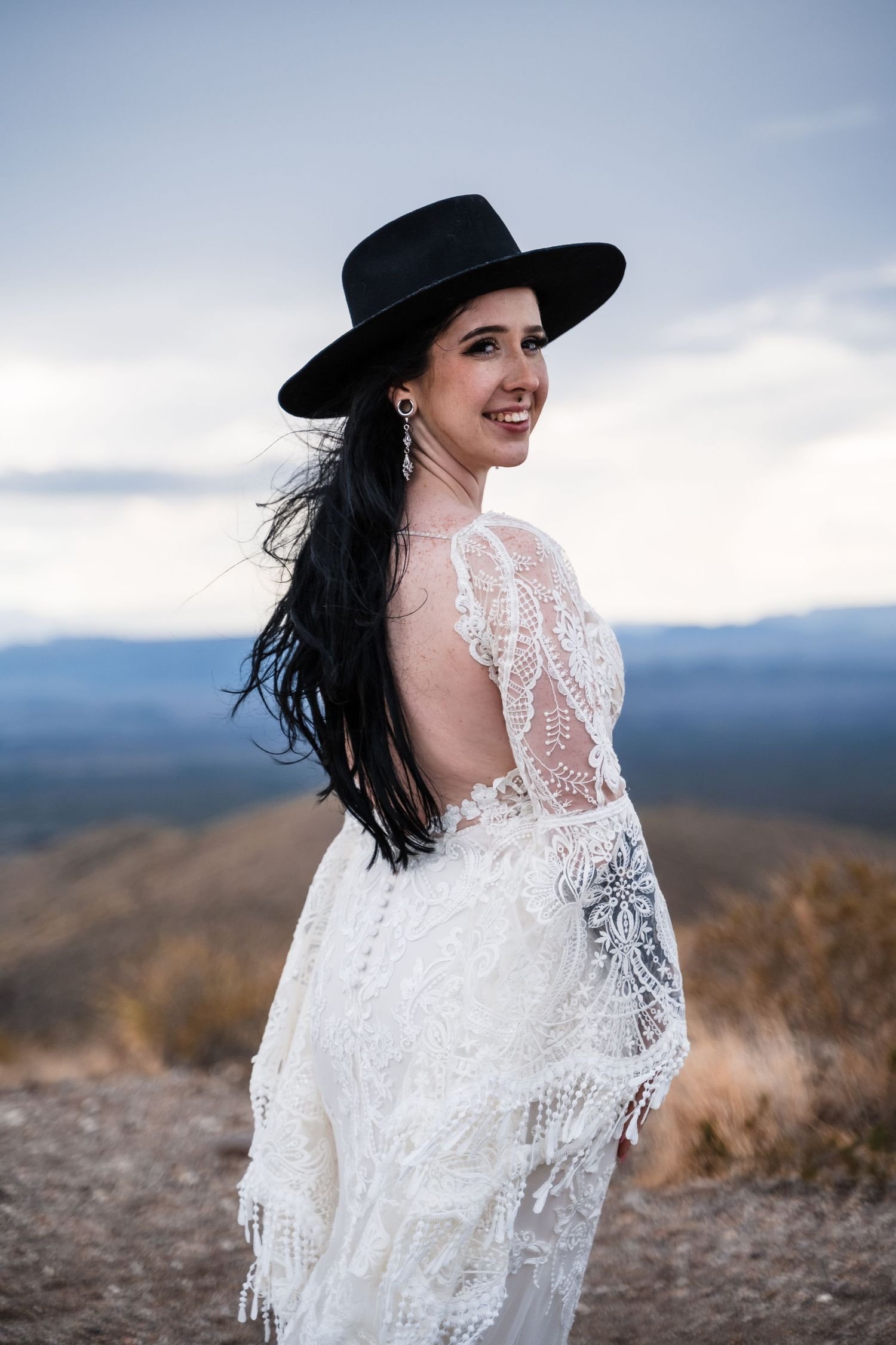 A woman in a white lace dress and black hat poses against a mountain landscape backdrop.