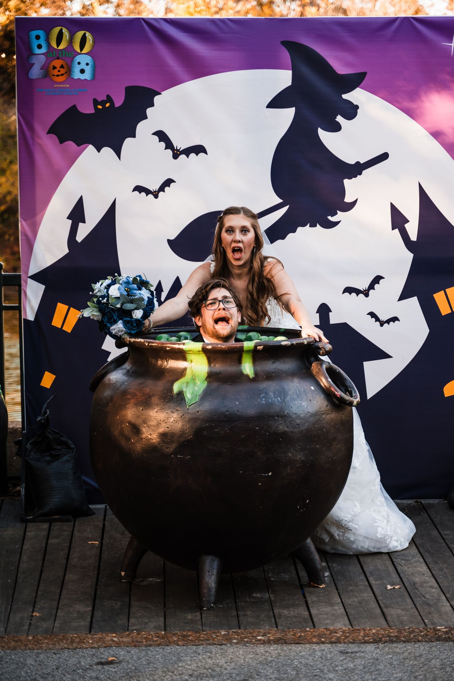 Fun Halloween-themed wedding photo with couple posing behind large cauldron prop.