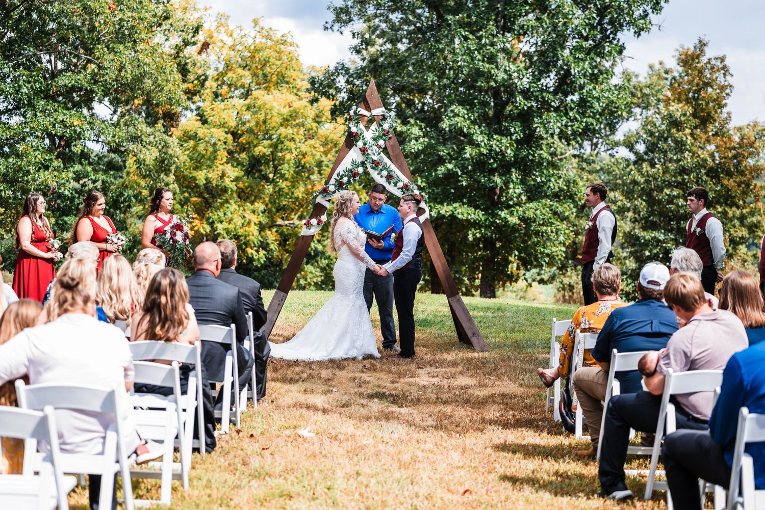 Outdoor wedding ceremony under trees with white chairs and guests watching the couple.