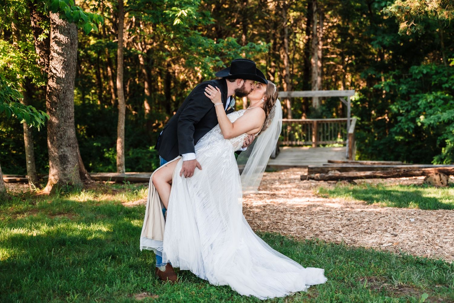 A couple shares a romantic dip pose in an outdoor garden setting with sunlit trees in the background.