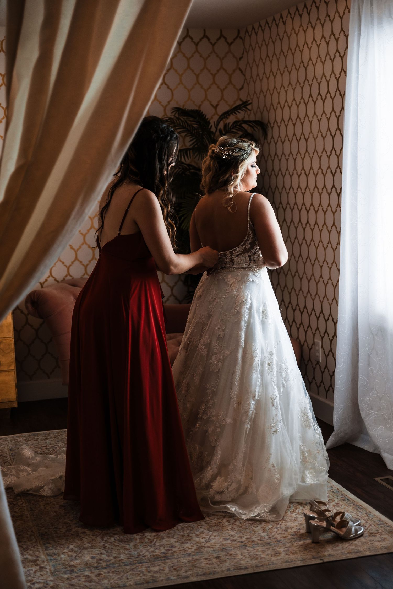 A bridesmaid in a burgundy dress helps with wedding preparations as she assists with a white lace gown.