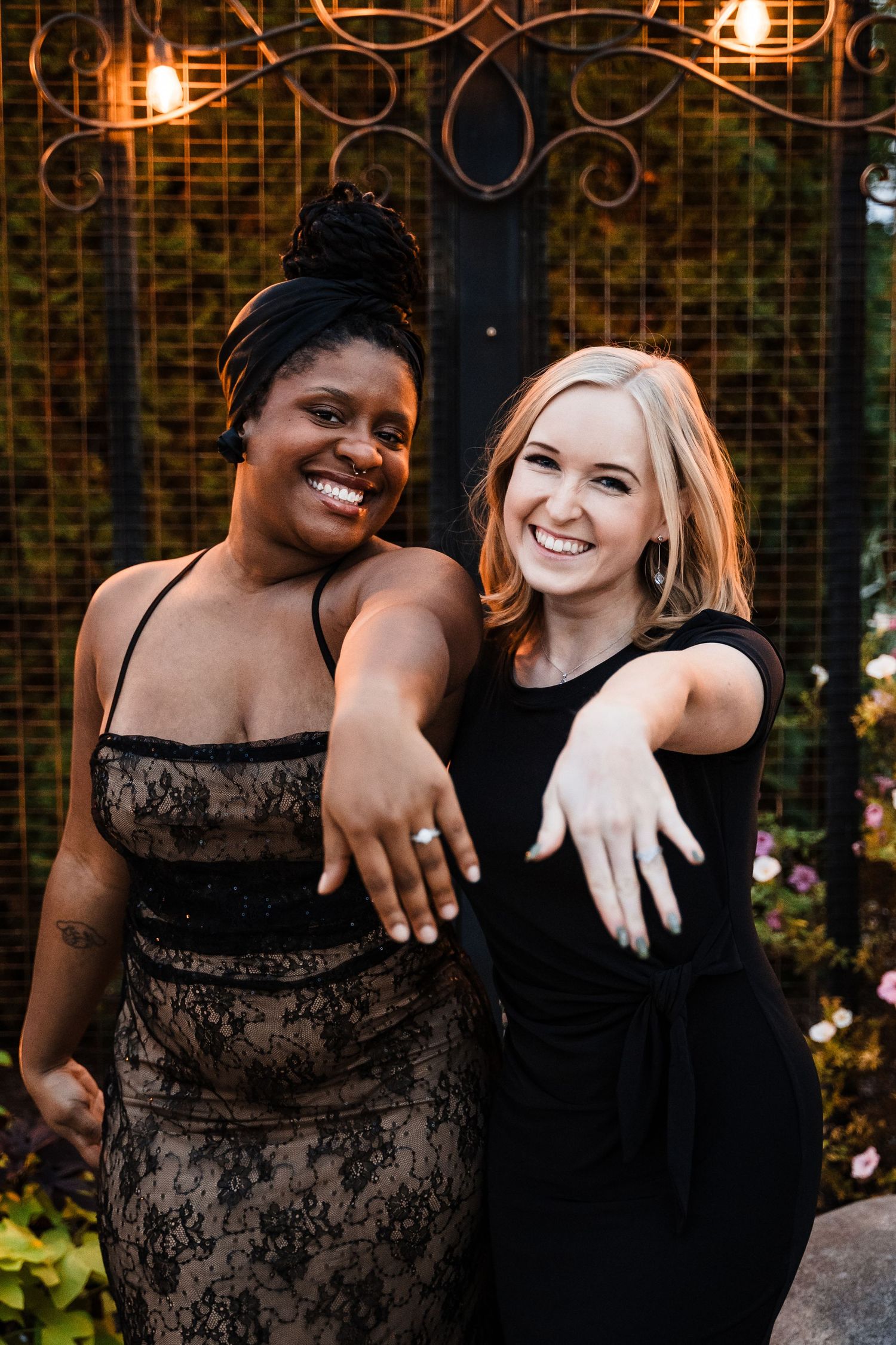 Two wedding guests in elegant black dresses showing off their rings in an outdoor garden setting at dusk.