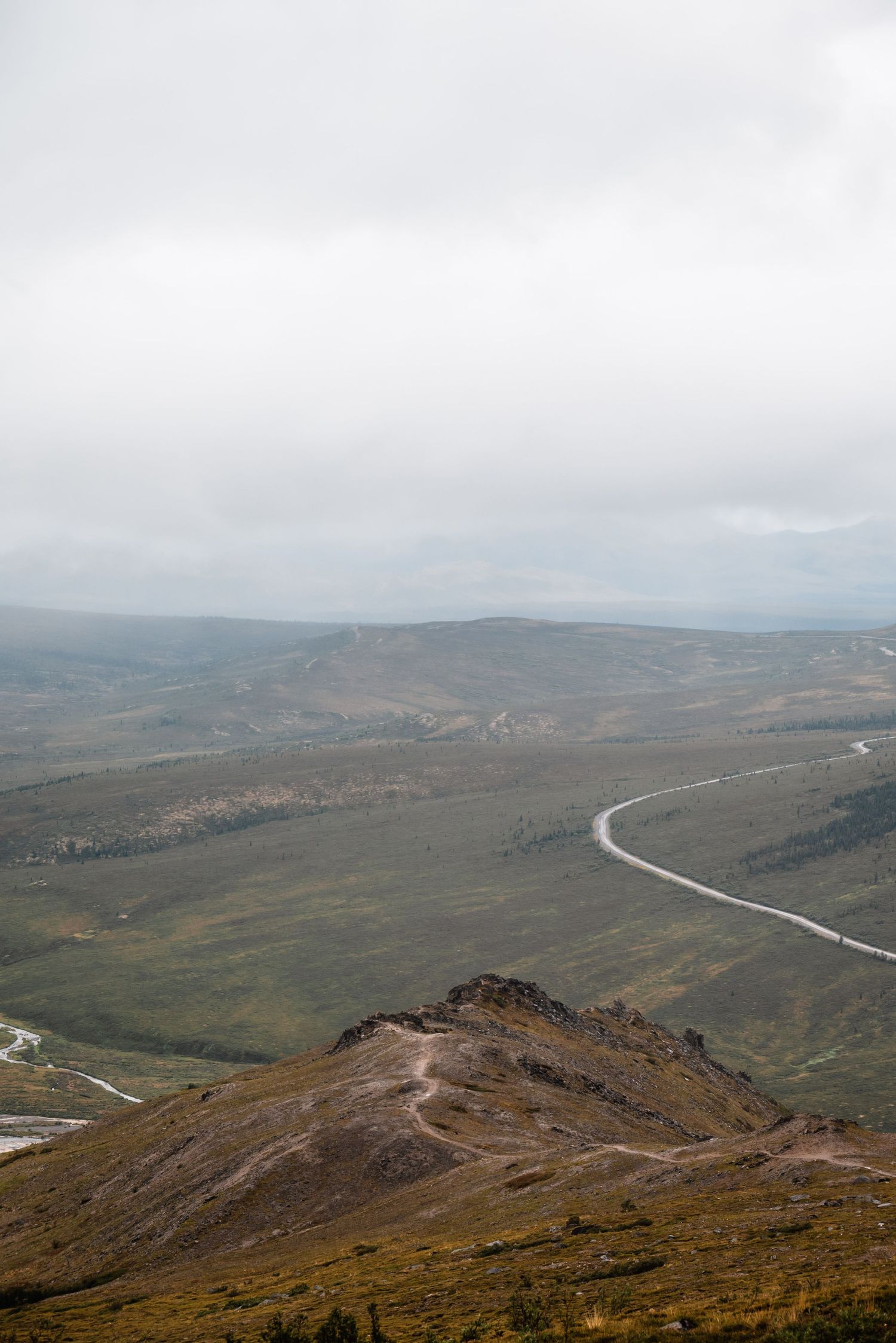 A winding mountain road curves through rugged terrain with distant hills and cloudy skies in the background.