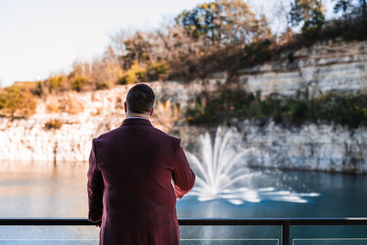 Person in burgundy suit watching fountain display in quarry lake at sunset.