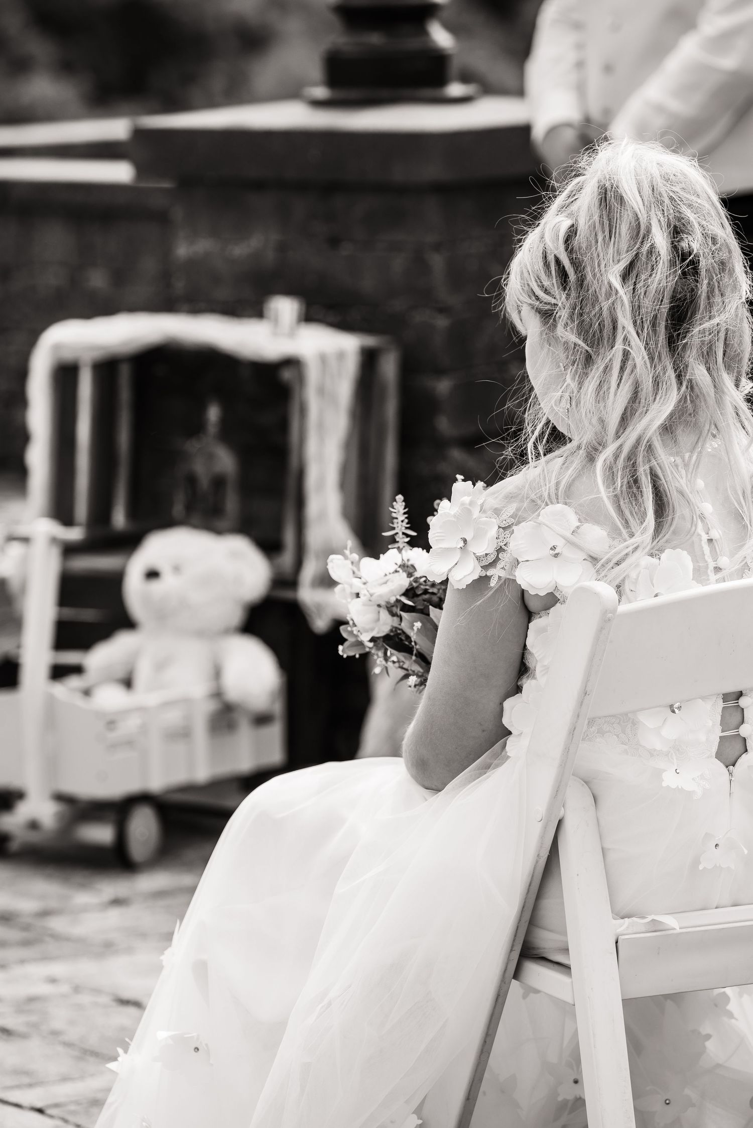 Black and white photo of someone in a wedding dress seated near a teddy bear.
