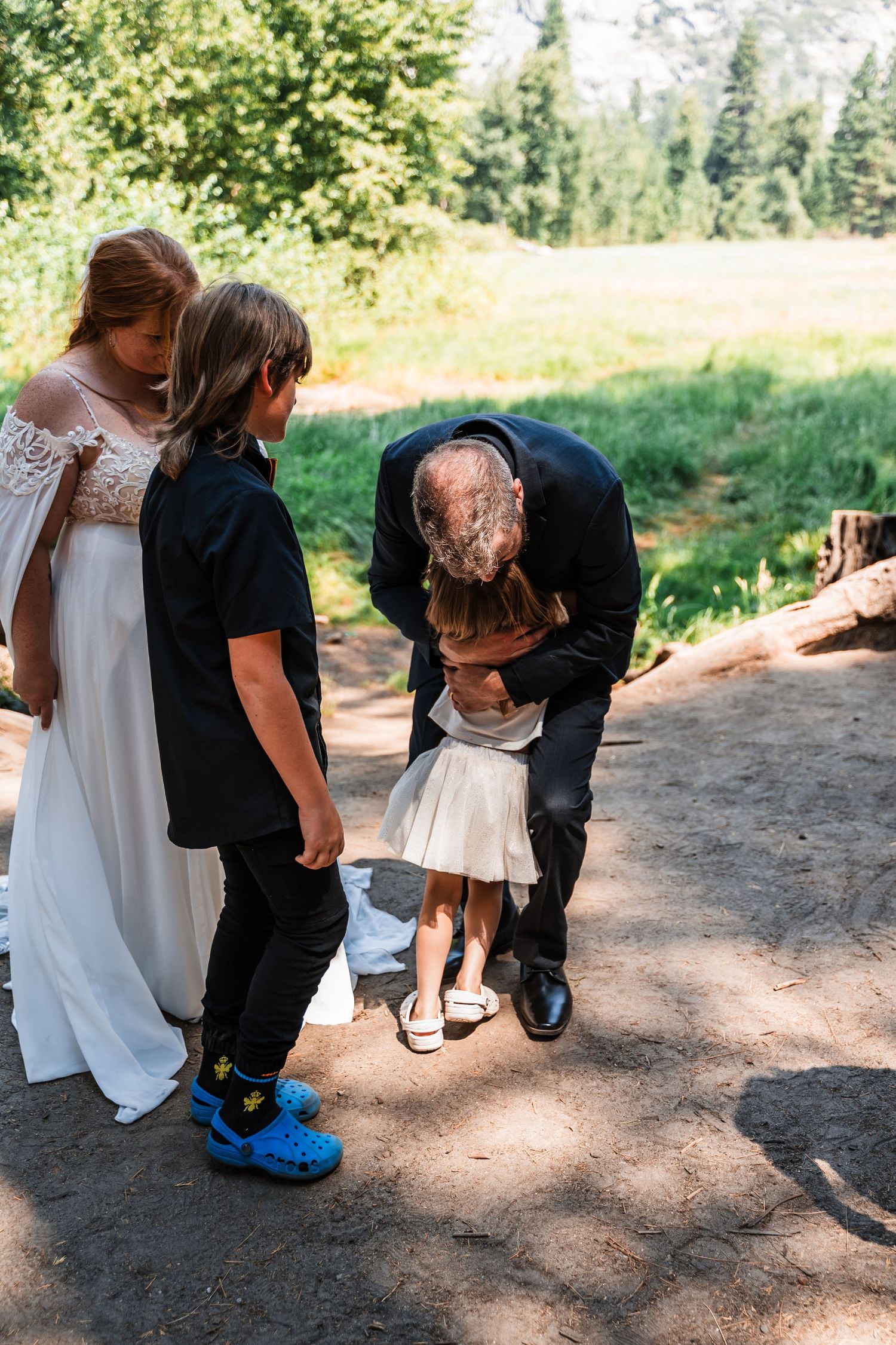 Family explores nature on a dirt path, kneeling and crouching together to examine something on the ground.