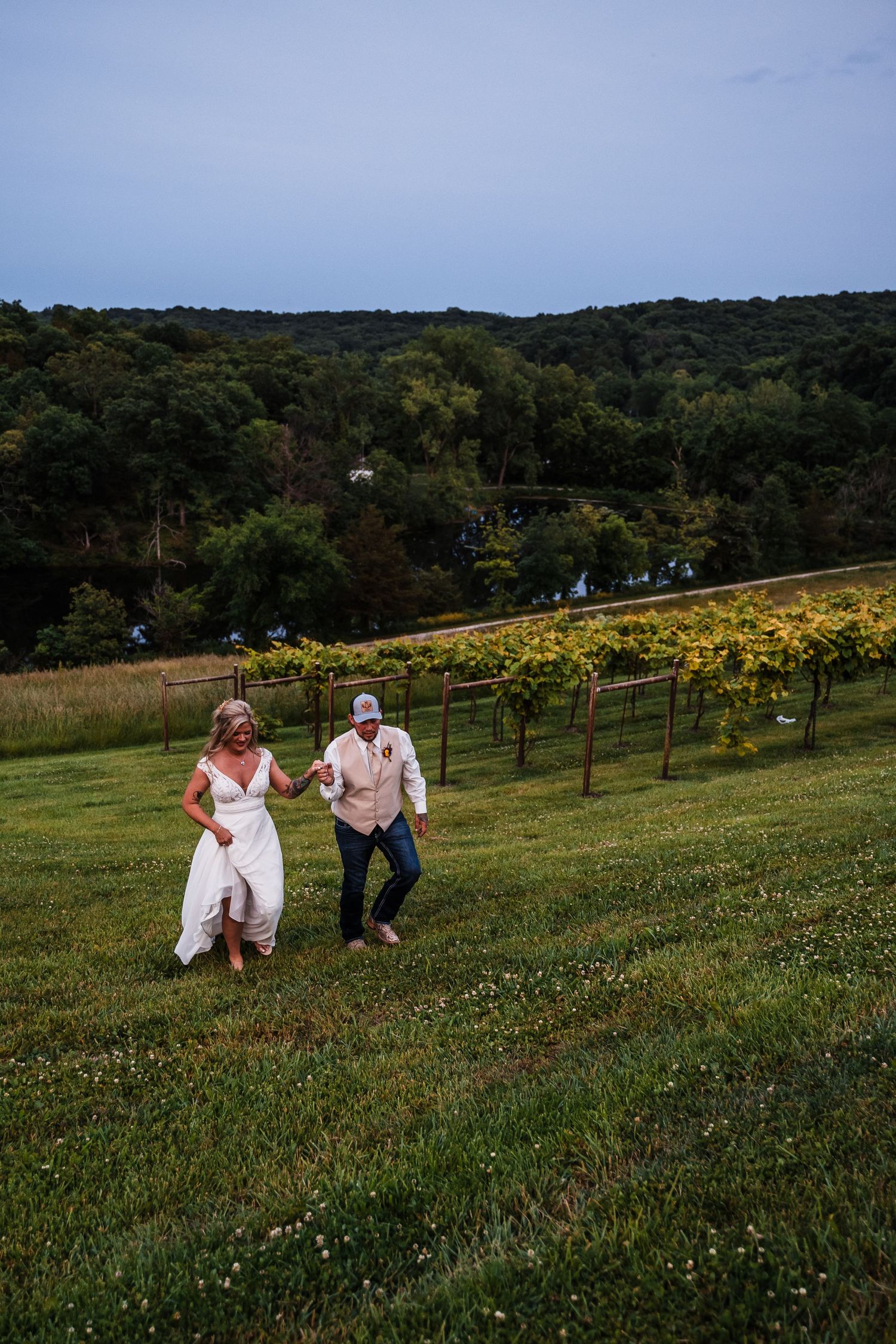 Couple poses playfully on grassy hillside with vineyard and mountains in background.