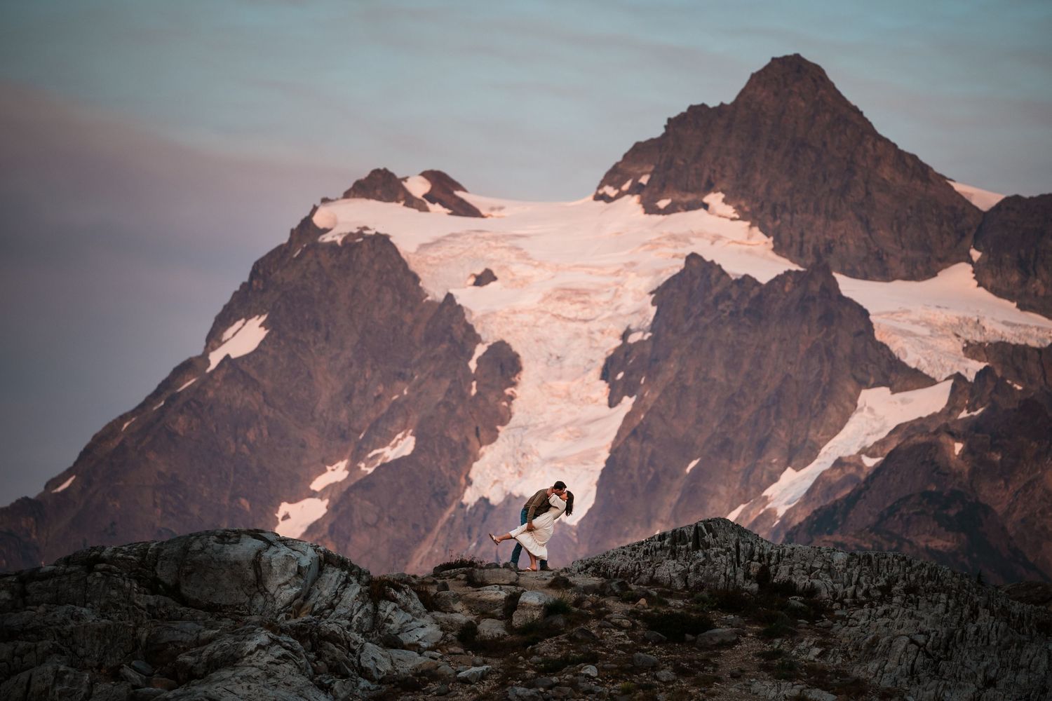 A panoramic mountain landscape with snow-capped peaks and rocky terrain against a cloudy sky at sunset.