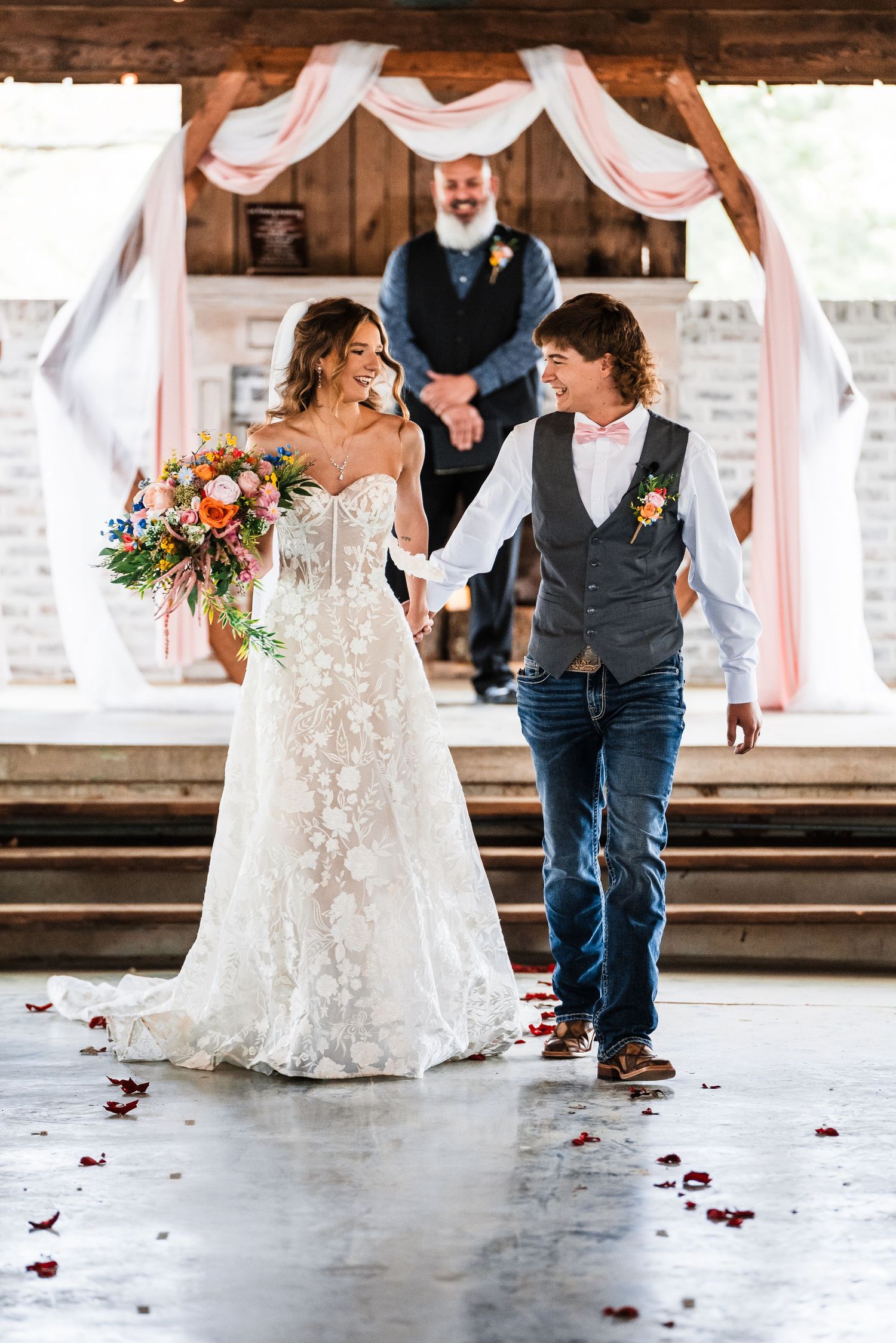 Newlyweds walk hand in hand under a rustic wooden arch decorated with white draping.