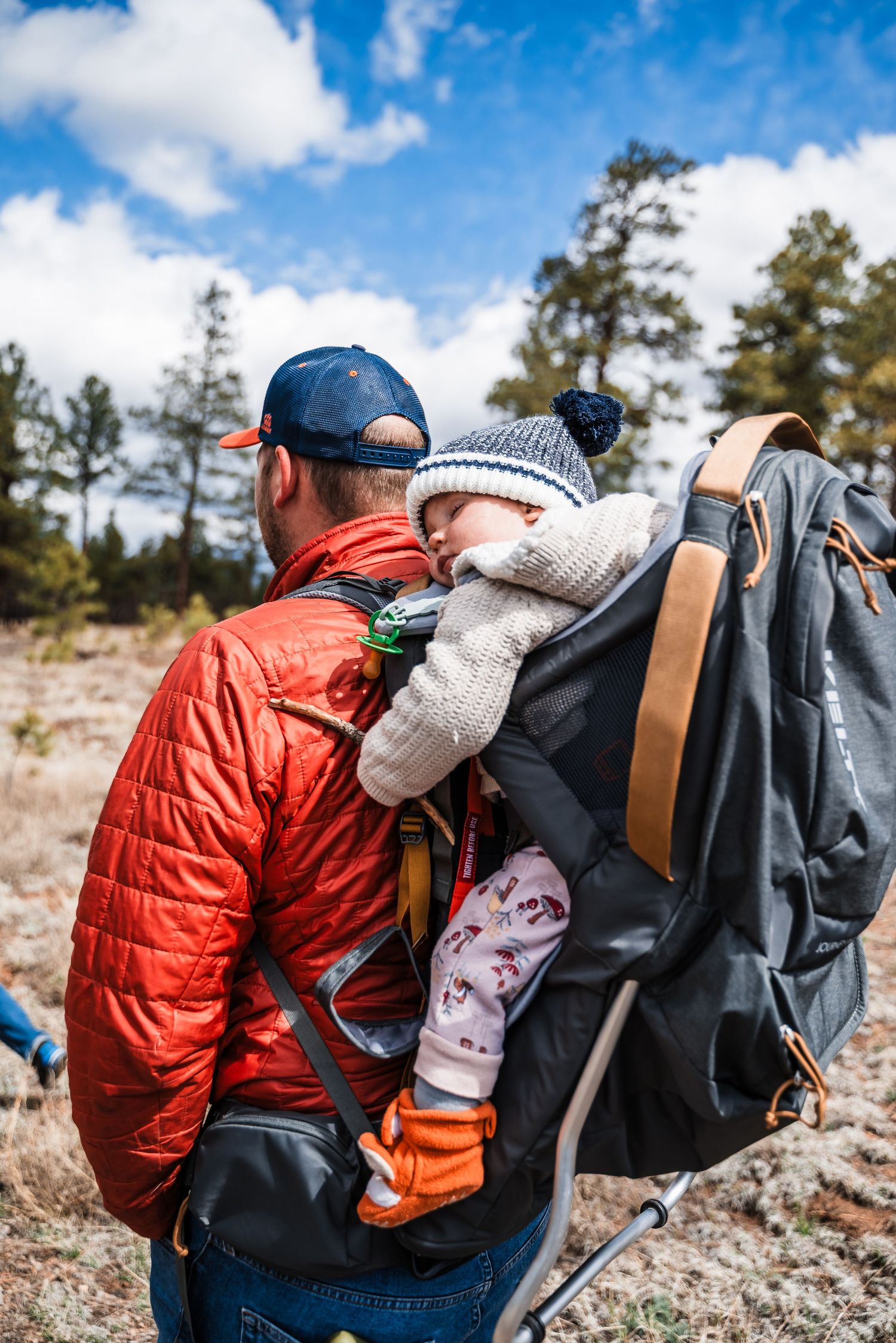 Hiker in orange jacket carrying backpack and hiking gear on a sunny mountain trail.