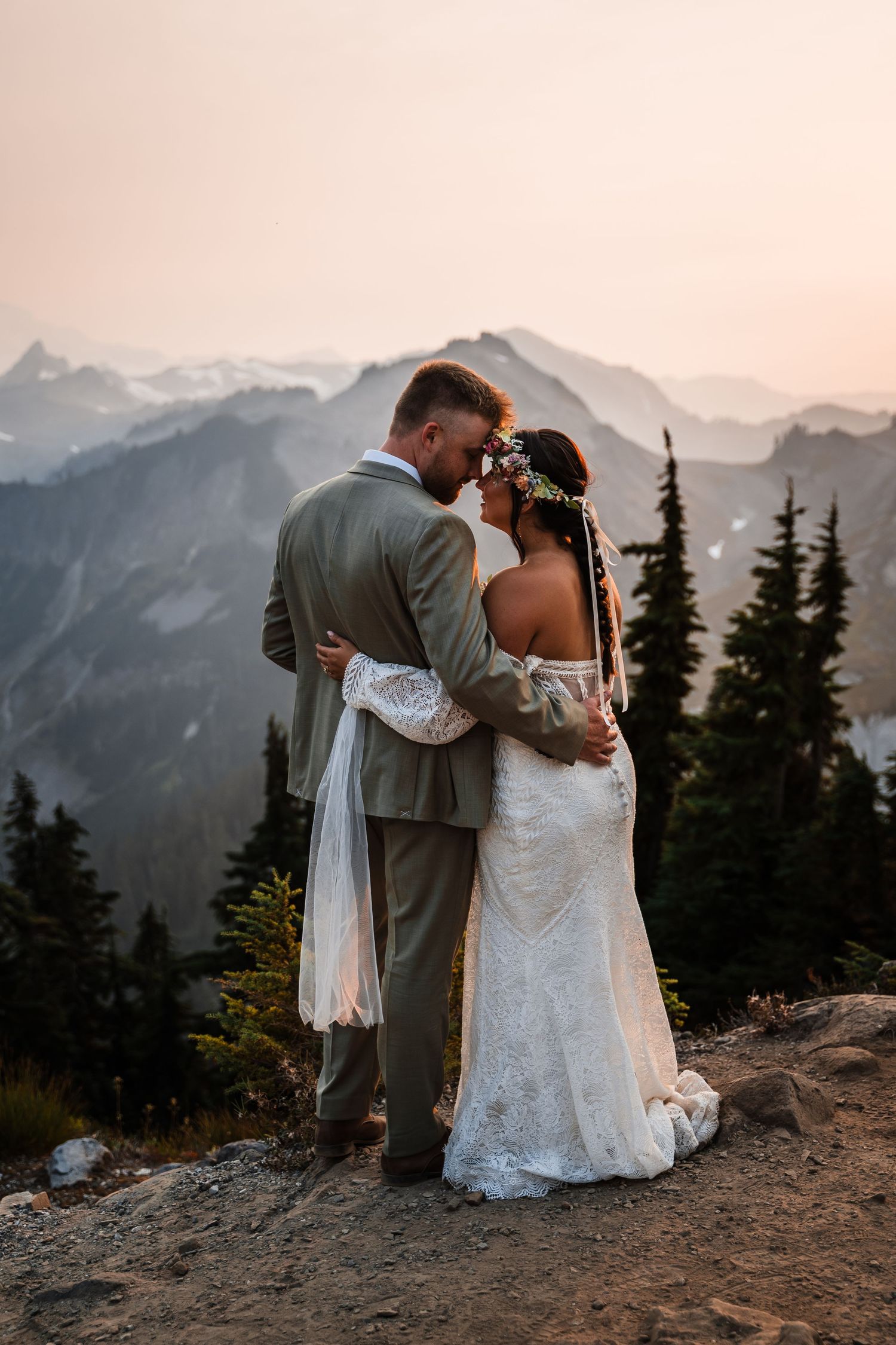 Newlyweds share an intimate moment on a mountain ridge with a misty sunset backdrop.