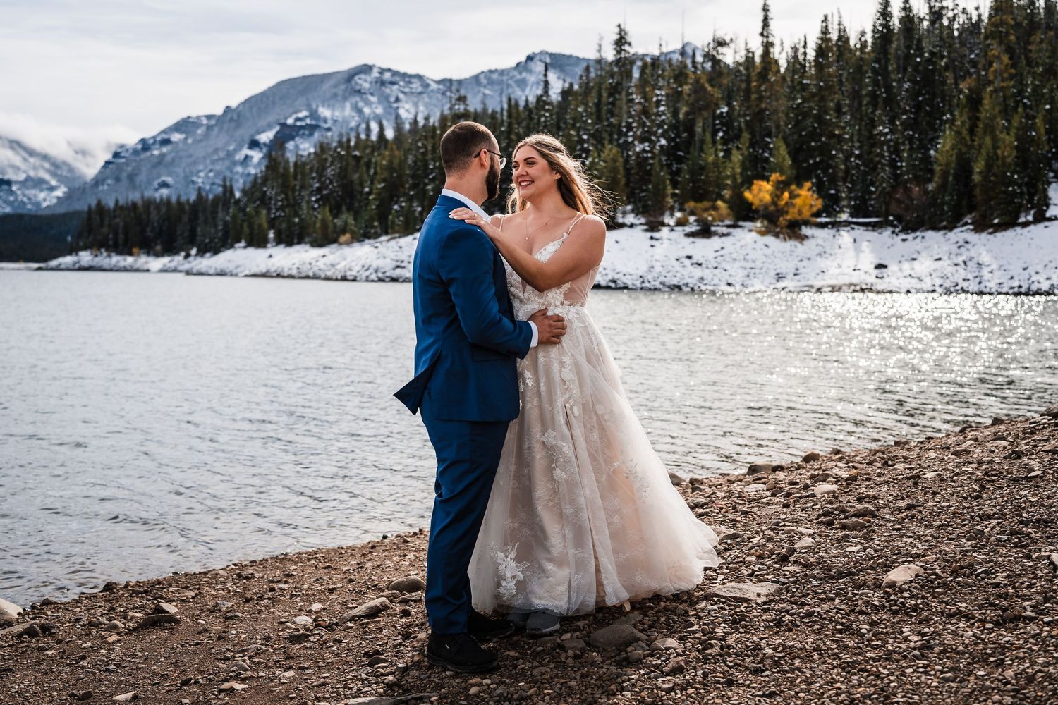 A winter wedding portrait by a snowy mountain lake with evergreen trees in the background.