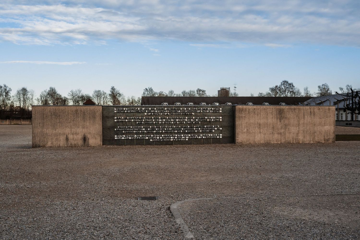 A concrete memorial wall with text stands alone in a barren landscape under cloudy skies.