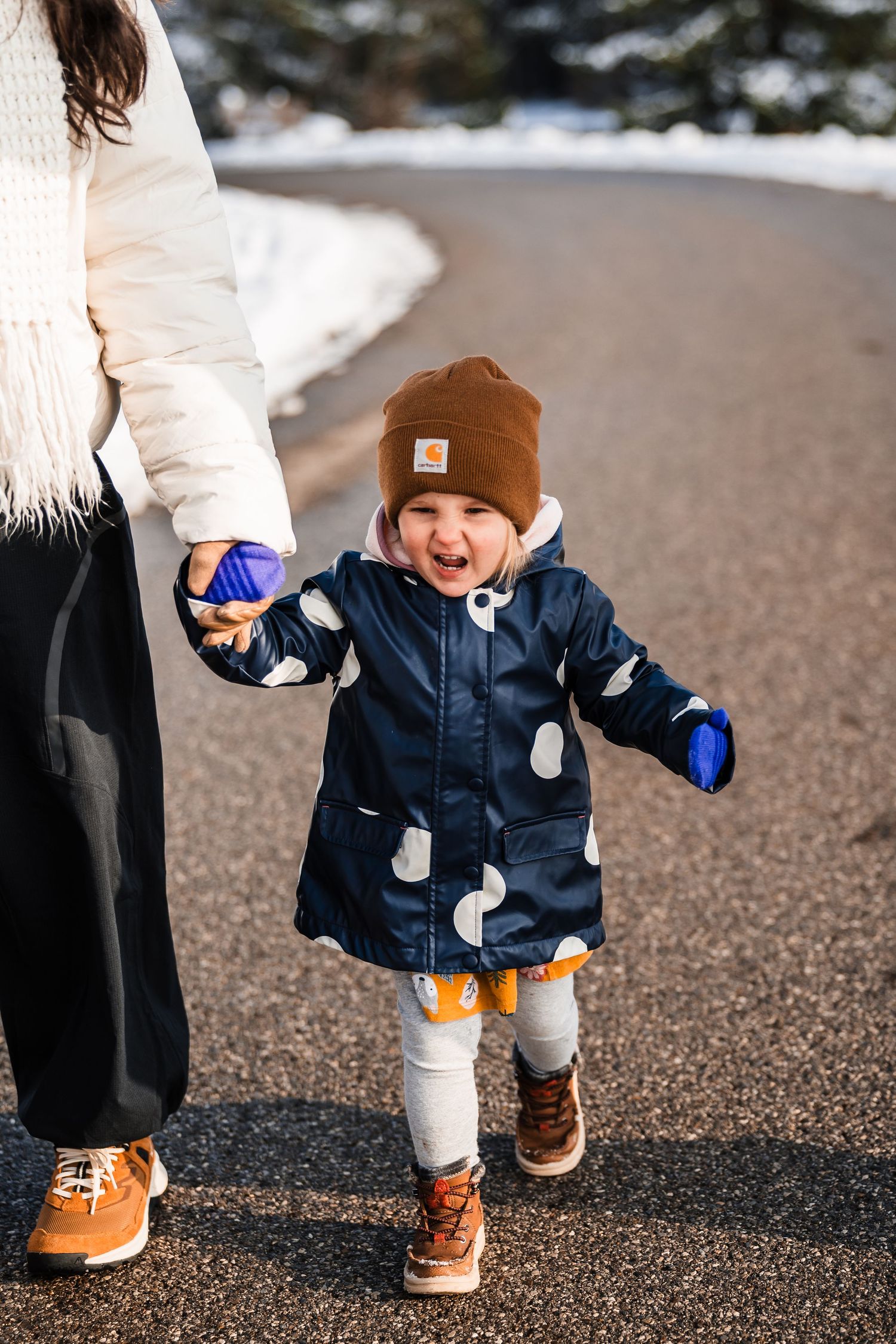Small child in navy coat and brown beanie takes steps on snowy path while wearing winter boots.