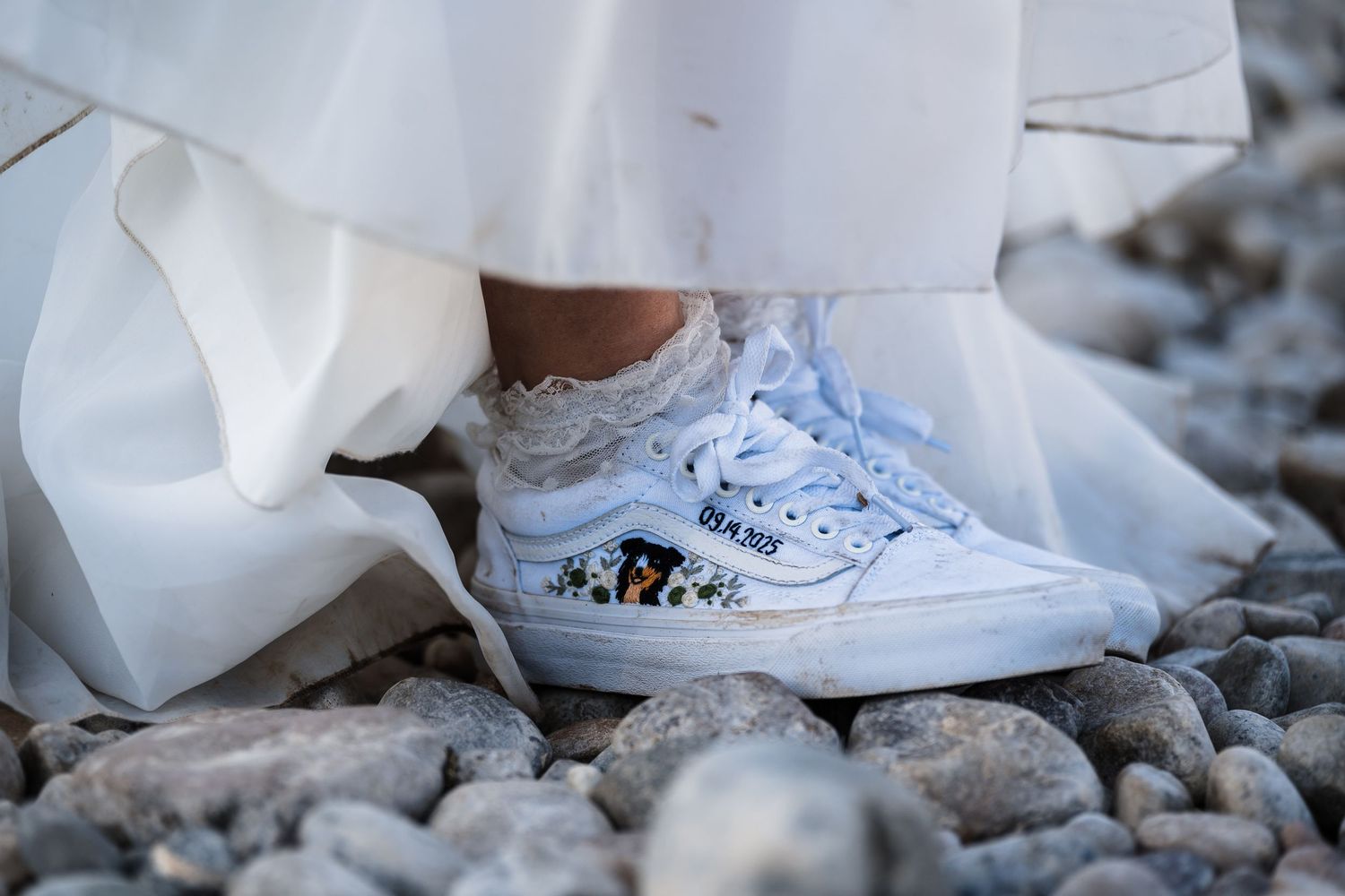 Close-up view of white wedding dress hem and sneakers on rocky ground showing casual bridal style.