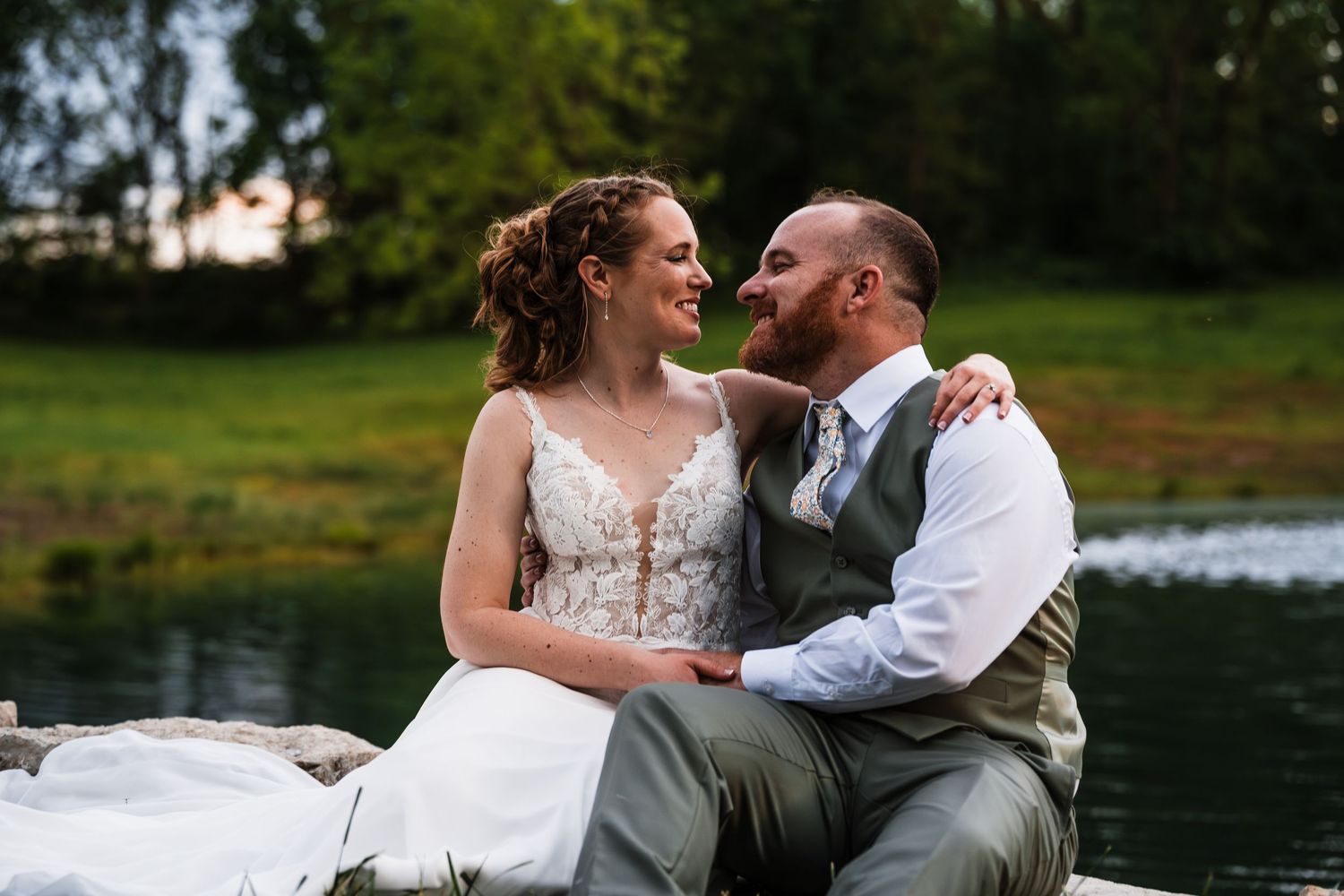Bride and groom sitting together by a peaceful lakeside, smiling at each other during golden hour.