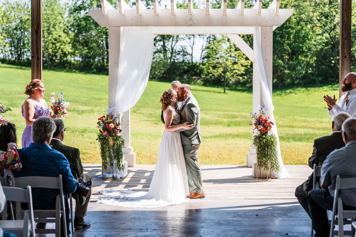 Bride and groom share their first kiss under a white draped arch adorned with colorful floral arrangements outdoors.
