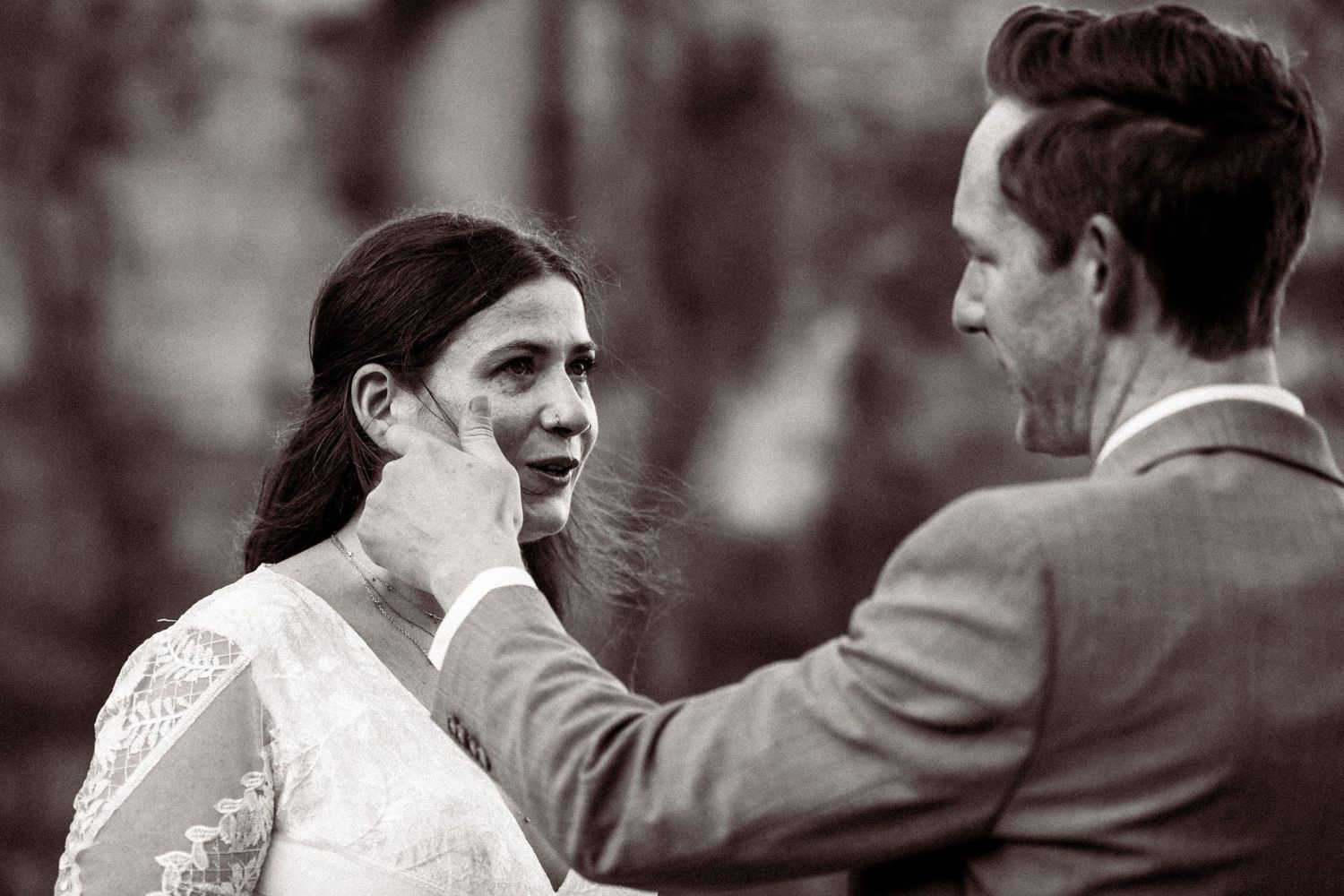 Groom tenderly wipes tears from bride's cheek during emotional outdoor wedding ceremony.