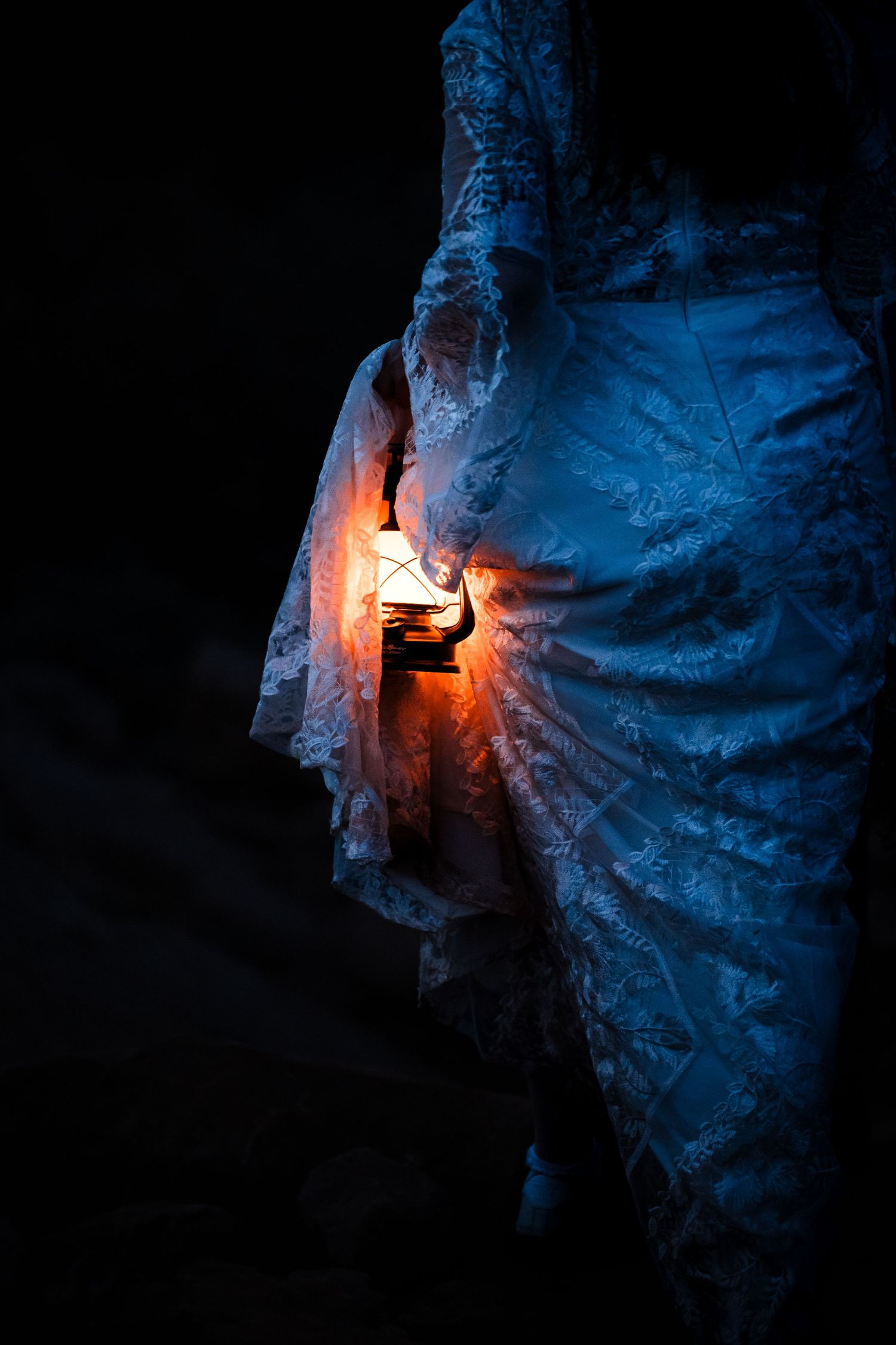 Bride in lace wedding gown holds glowing lantern in dramatic dark canyon setting.