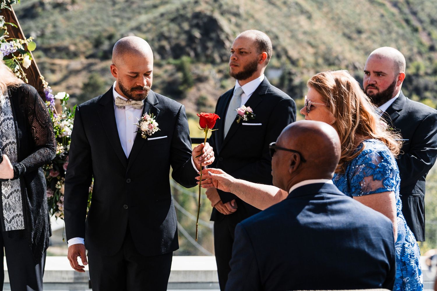 Groom in grey suit receiving red rose from guest during outdoor mountain wedding ceremony.