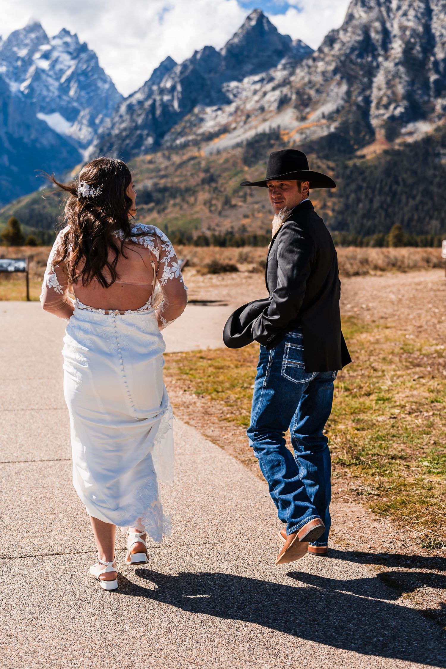 A couple in western attire walking on a path with snow-capped Grand Teton mountains in the background.