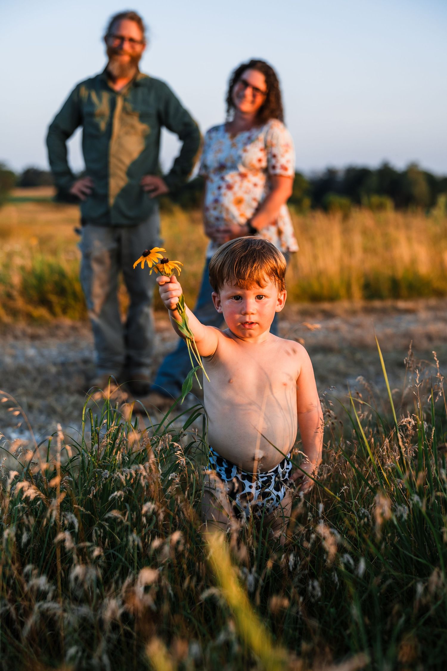 A young child explores a field of wildflowers at sunset while two people watch in the background.