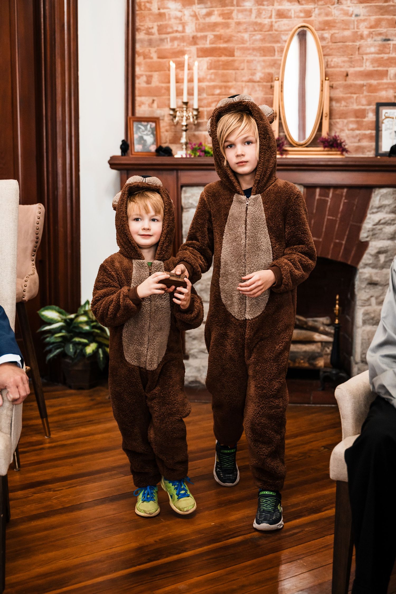 Two children wearing matching brown bear costumes stand in a cozy living room with a brick fireplace.