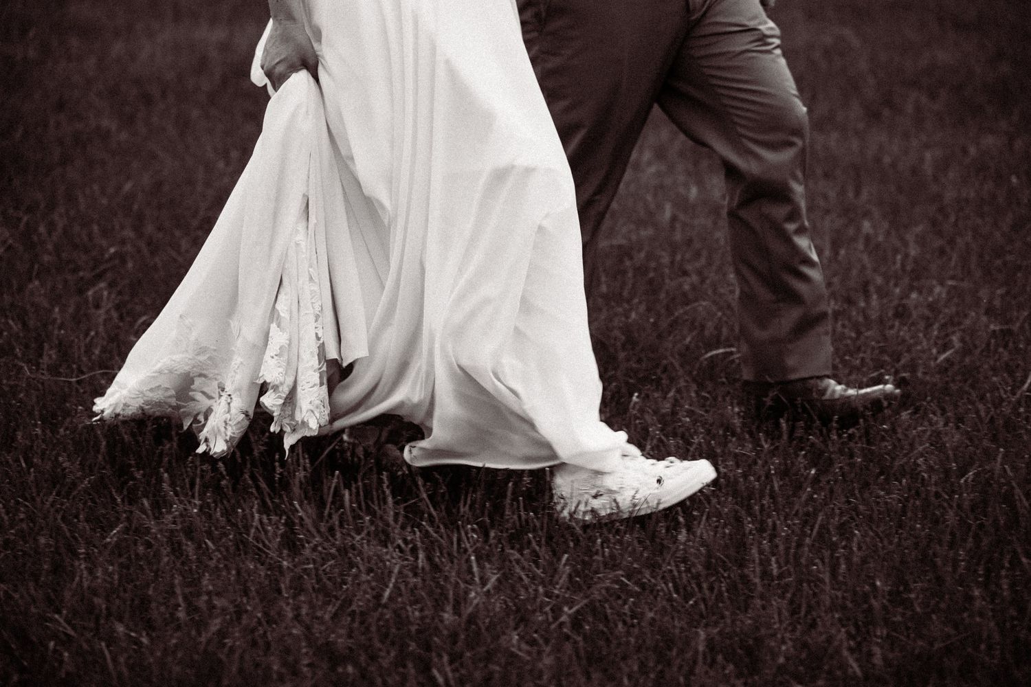 Black and white close-up of bride in lace gown wearing white sneakers walking with groom across grass.