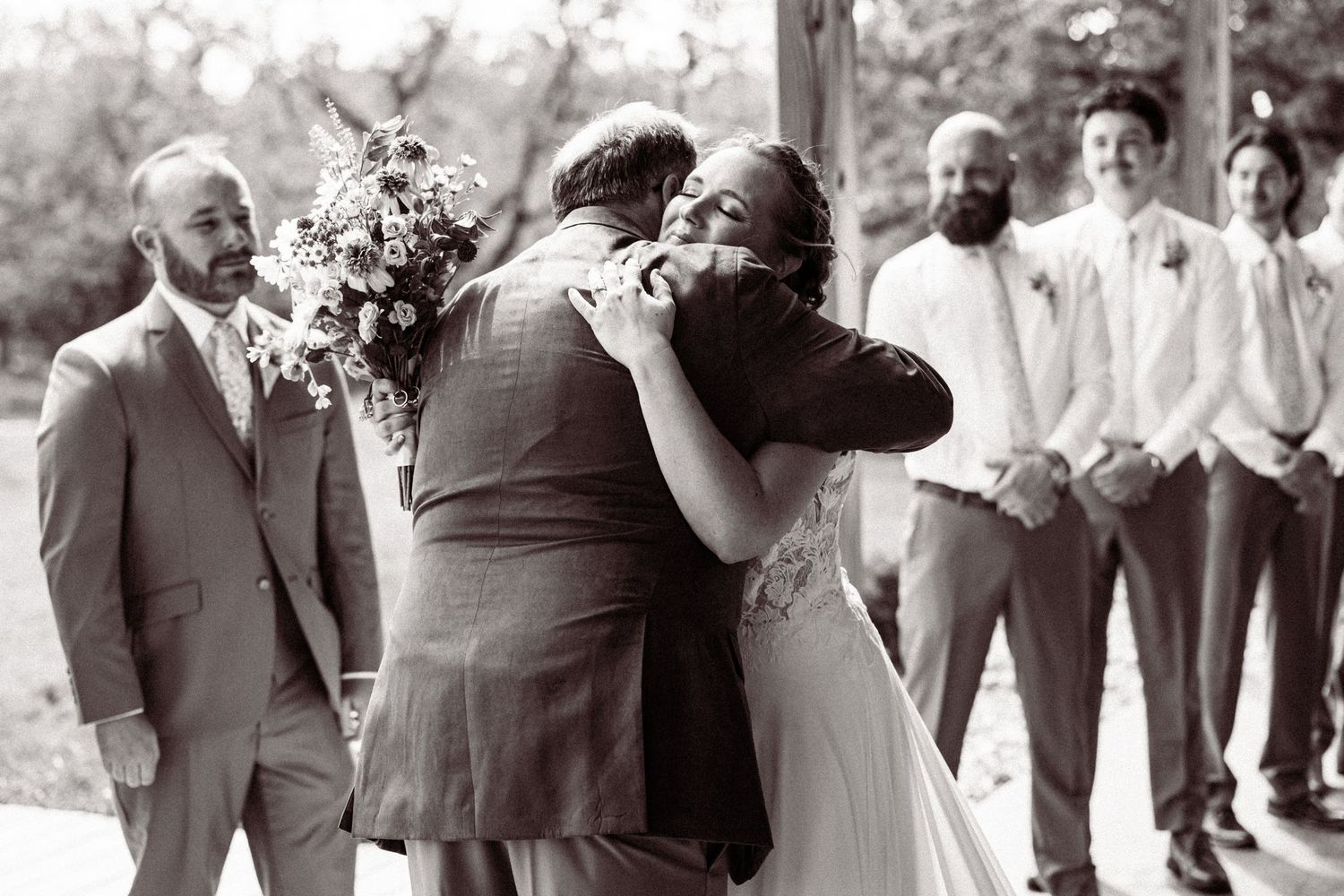 Bride embraces a family member in an emotional black and white wedding ceremony moment with groomsmen watching.