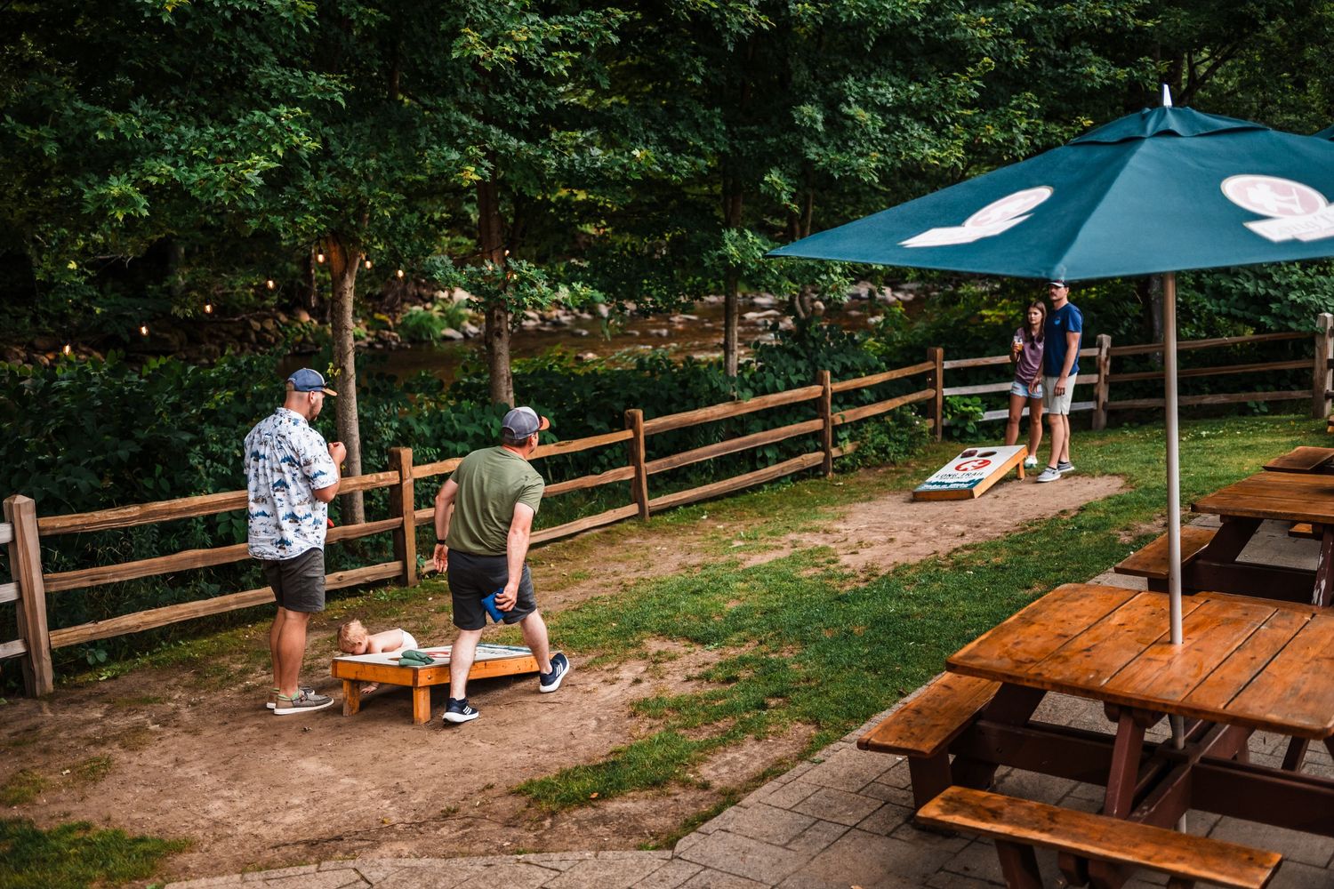 People enjoying an outdoor picnic area with wooden benches and a blue umbrella near a rustic fence on a summer day.