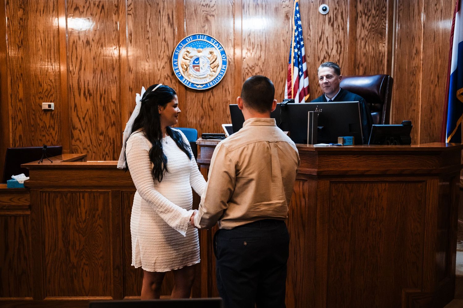 A couple stands before a judge in a courtroom with wood-paneled walls and the official state seal.