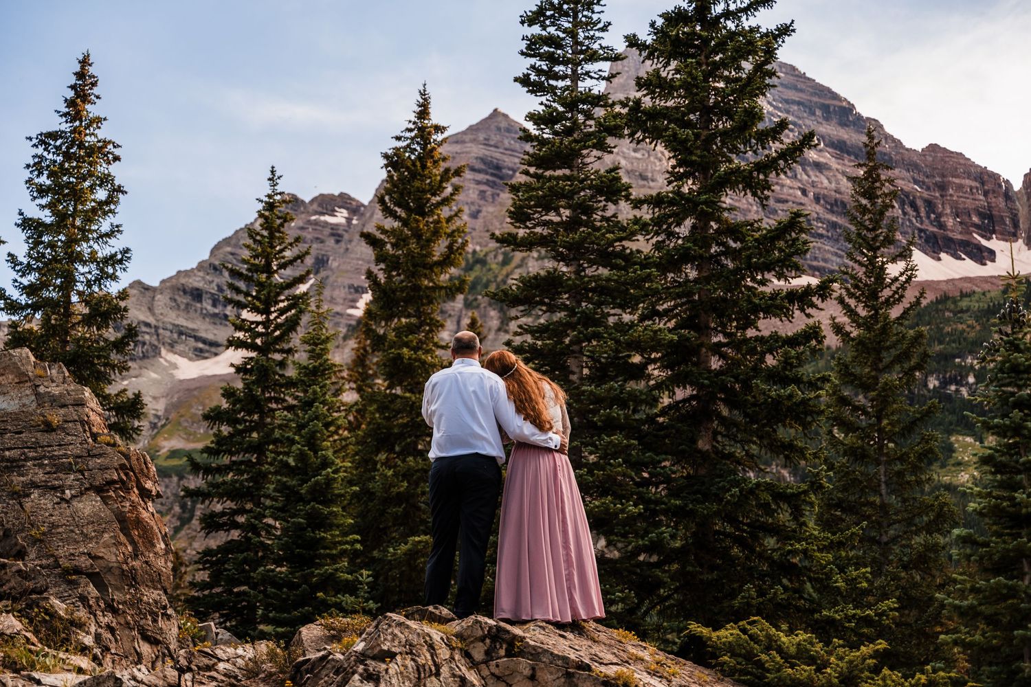 A couple embraces while overlooking majestic mountain peaks and evergreen forests.