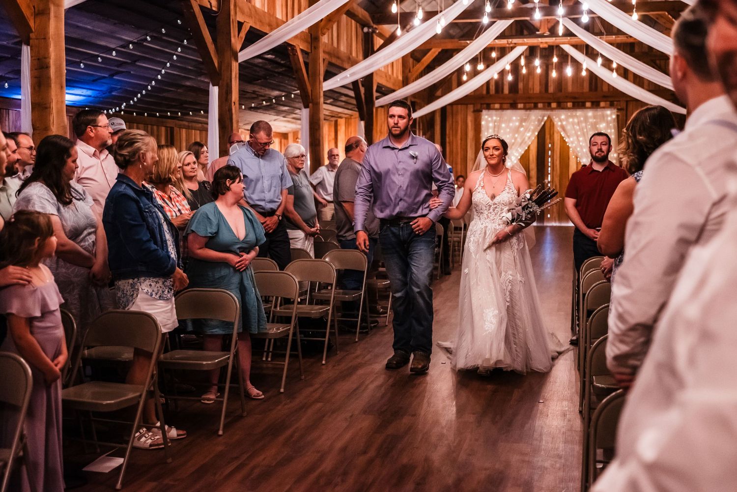 Wedding guests watch a couple enter their barn reception venue with twinkle lights and wooden beams overhead.