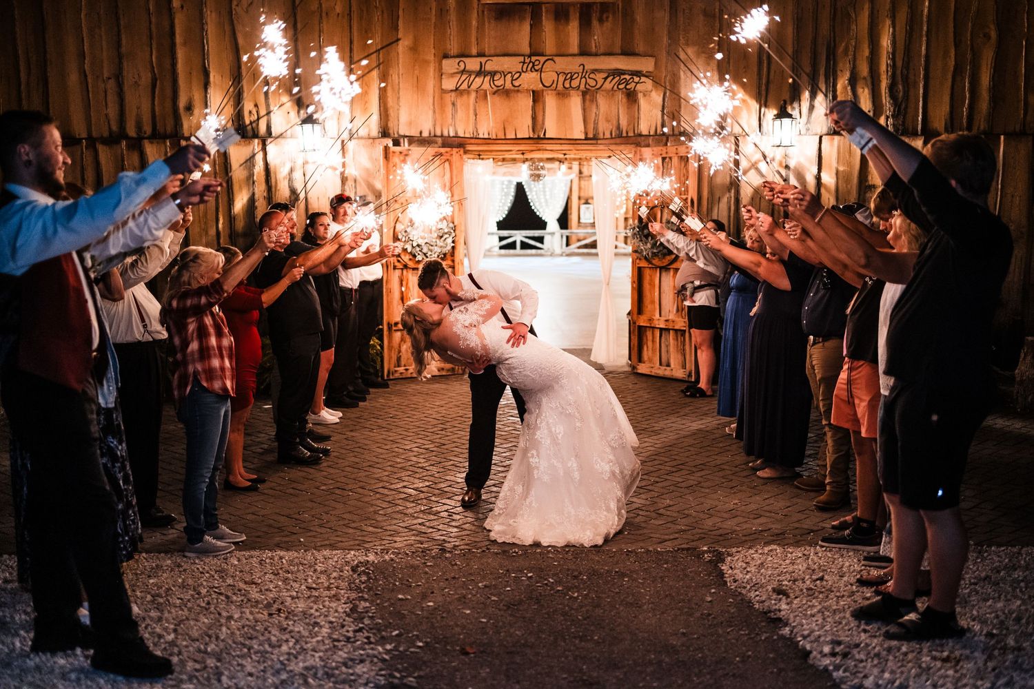 Newlyweds share romantic kiss under sparkler exit tunnel at rustic barn wedding reception.