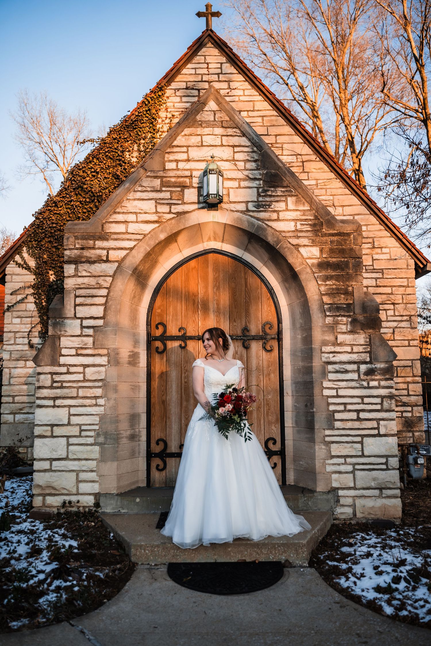 A bride in a white gown holds a bouquet while standing in front of a stone chapel entrance with snow on the ground at sunset.