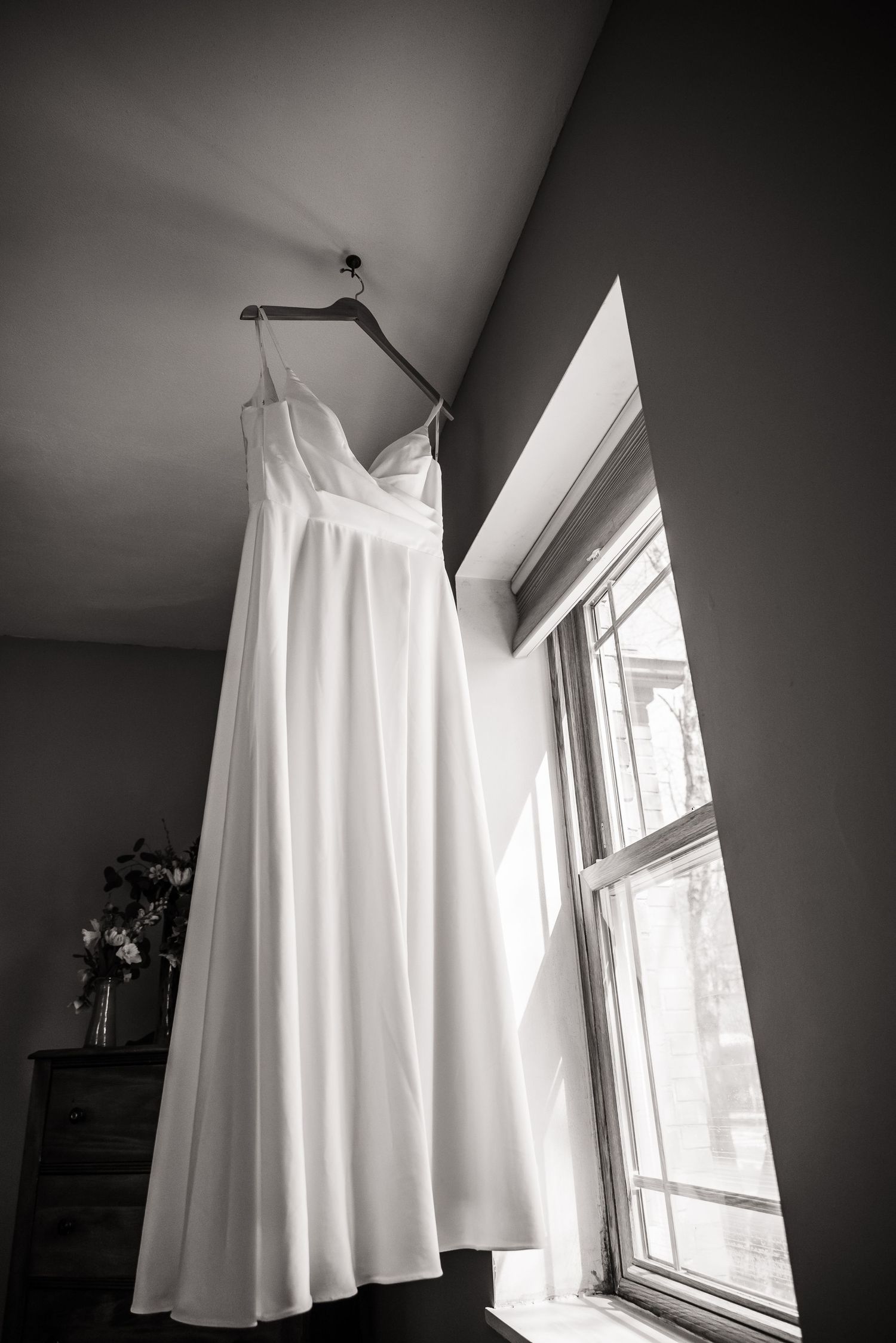 A white wedding dress hanging by a window in dramatic black and white photography.