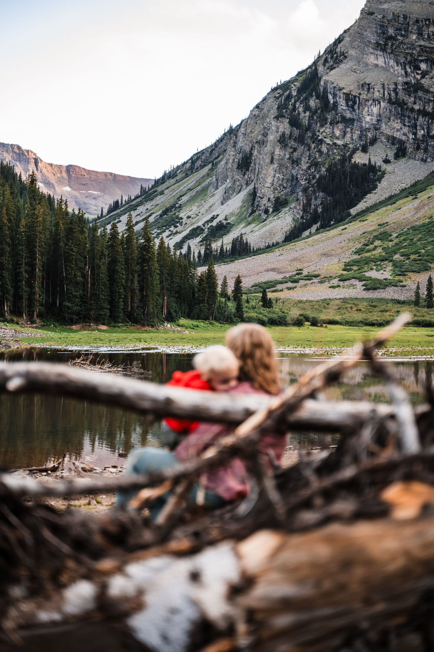 Serene mountain lake reflects rugged peaks and evergreen forest while camping gear rests on fallen logs in the foreground.