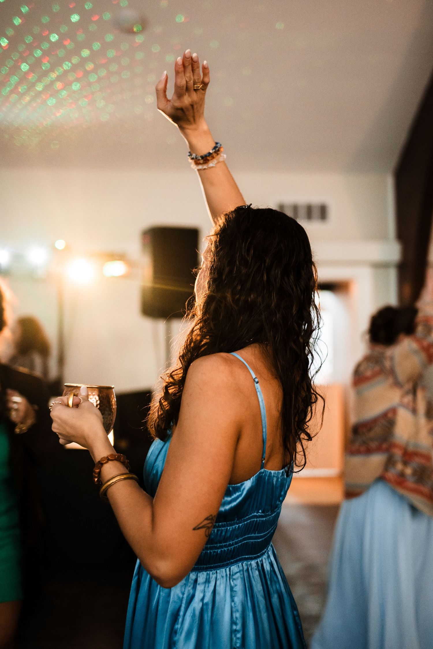 A person in a blue dress raises their hand and dances under sparkly lights at a celebration.