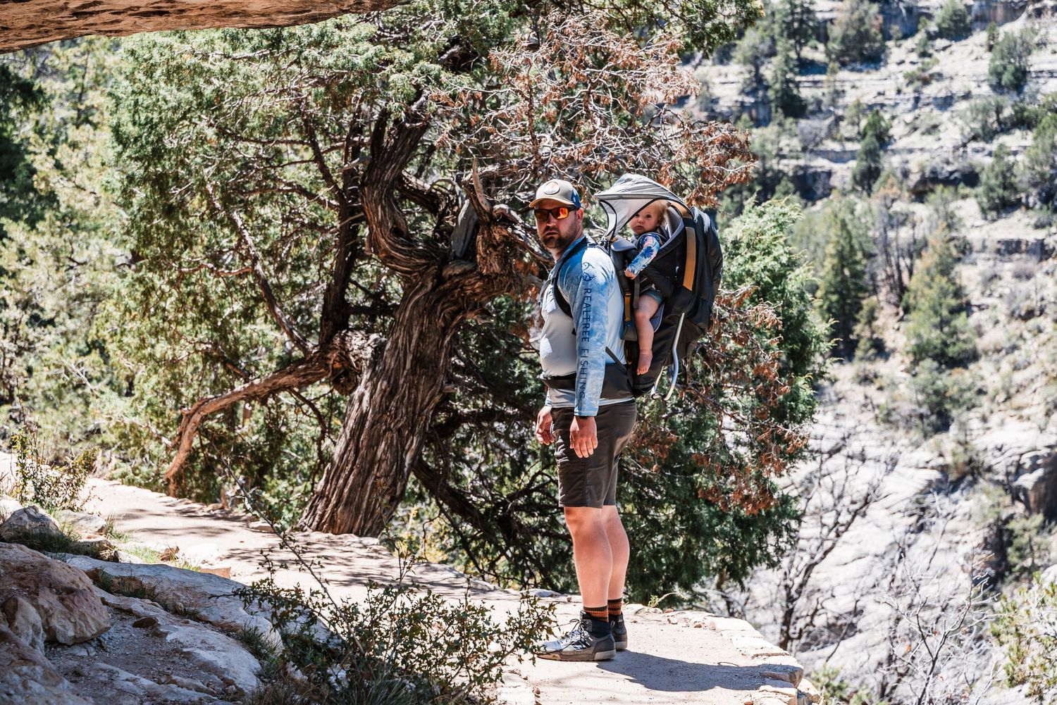 A hiker stands on a rocky trail surrounded by juniper trees and mountain scenery.