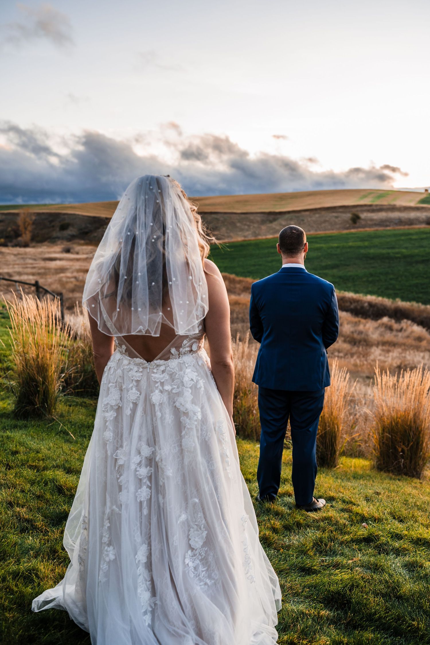 Bride in flowing white dress and groom in navy suit walk through scenic countryside at sunset.