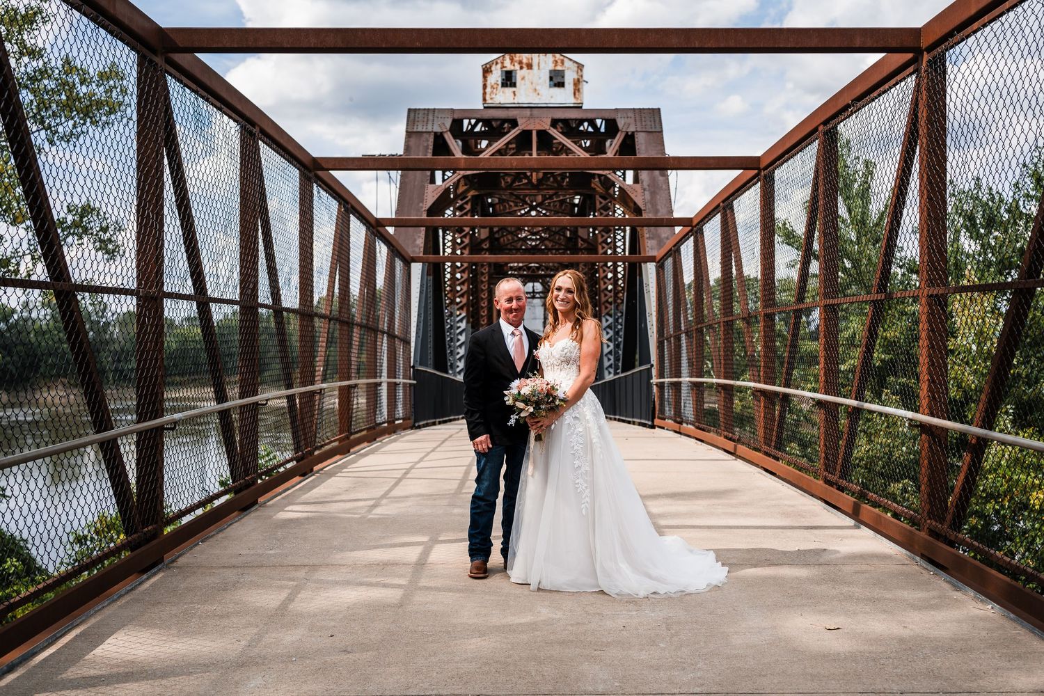 A couple poses on a rustic wooden bridge surrounded by lush greenery and natural scenery.