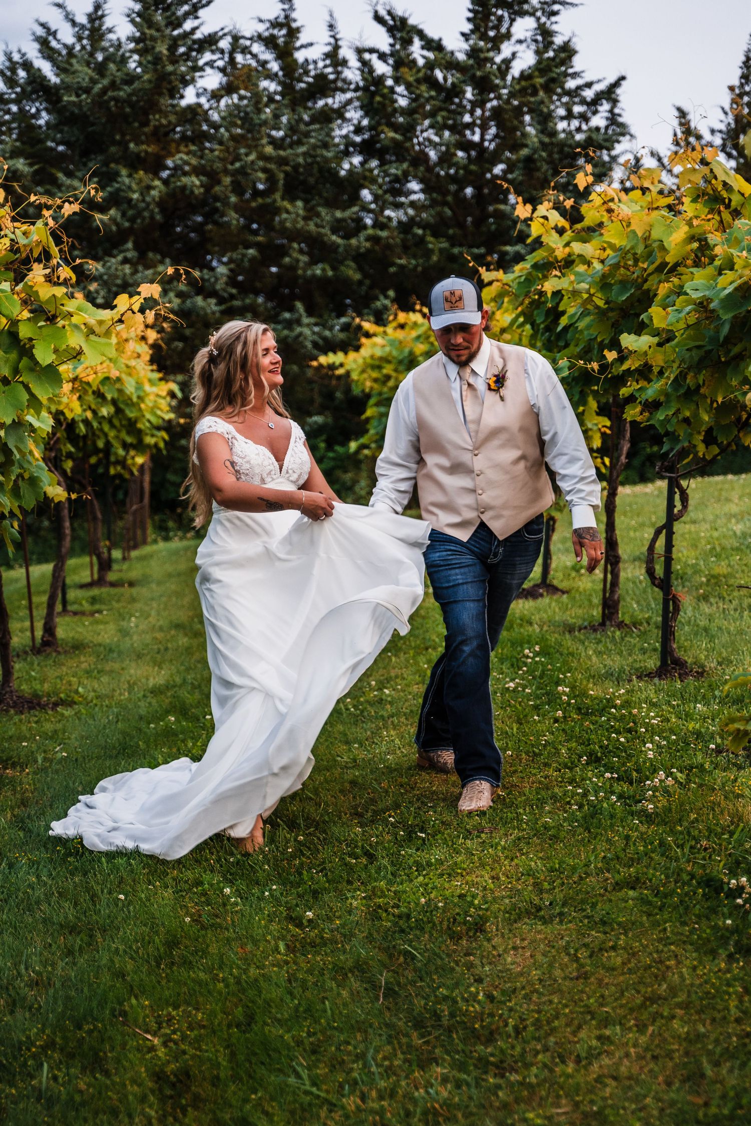 A couple strolls through a vineyard, with the bride's white dress flowing in the breeze against green grape vines.