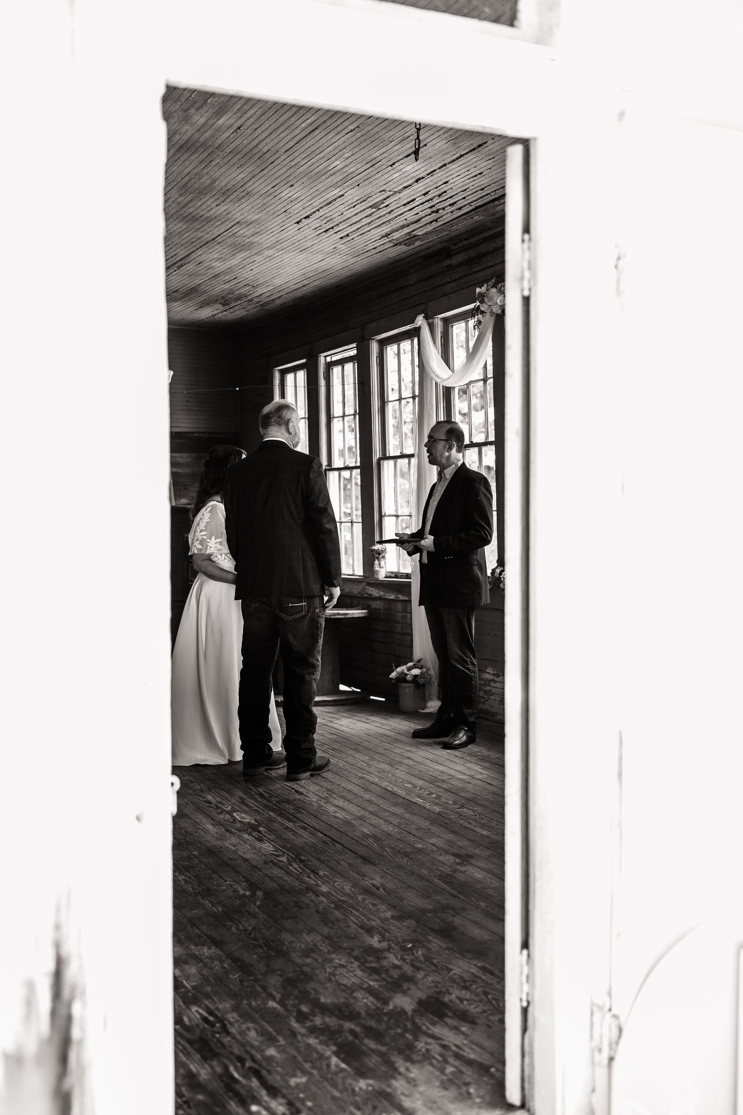 Black and white photo showing wedding ceremony inside rustic wooden room with tall windows.