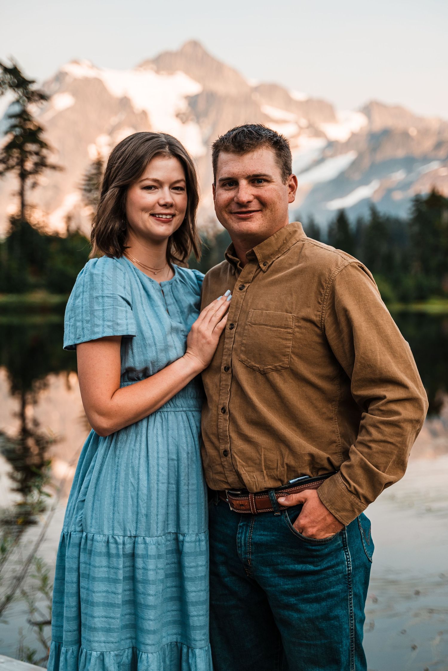 A couple poses together outdoors at sunset with mountains and trees visible in the background.
