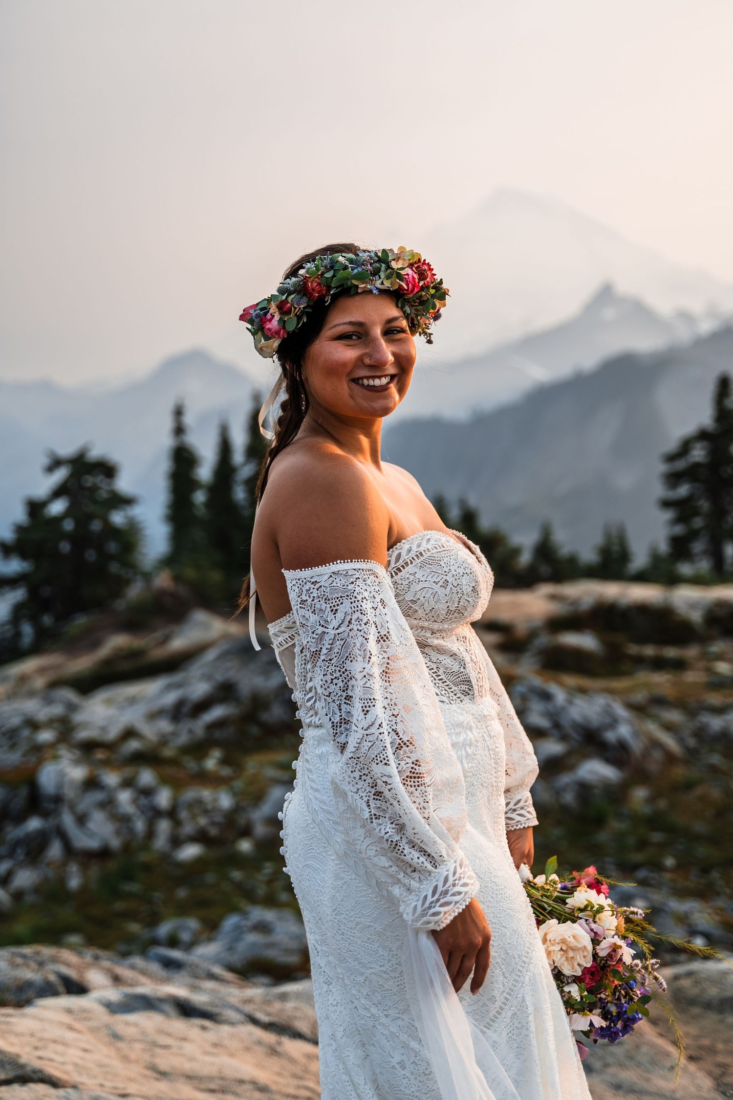 A bride in a lace dress and floral crown poses against a mountain backdrop at sunset.