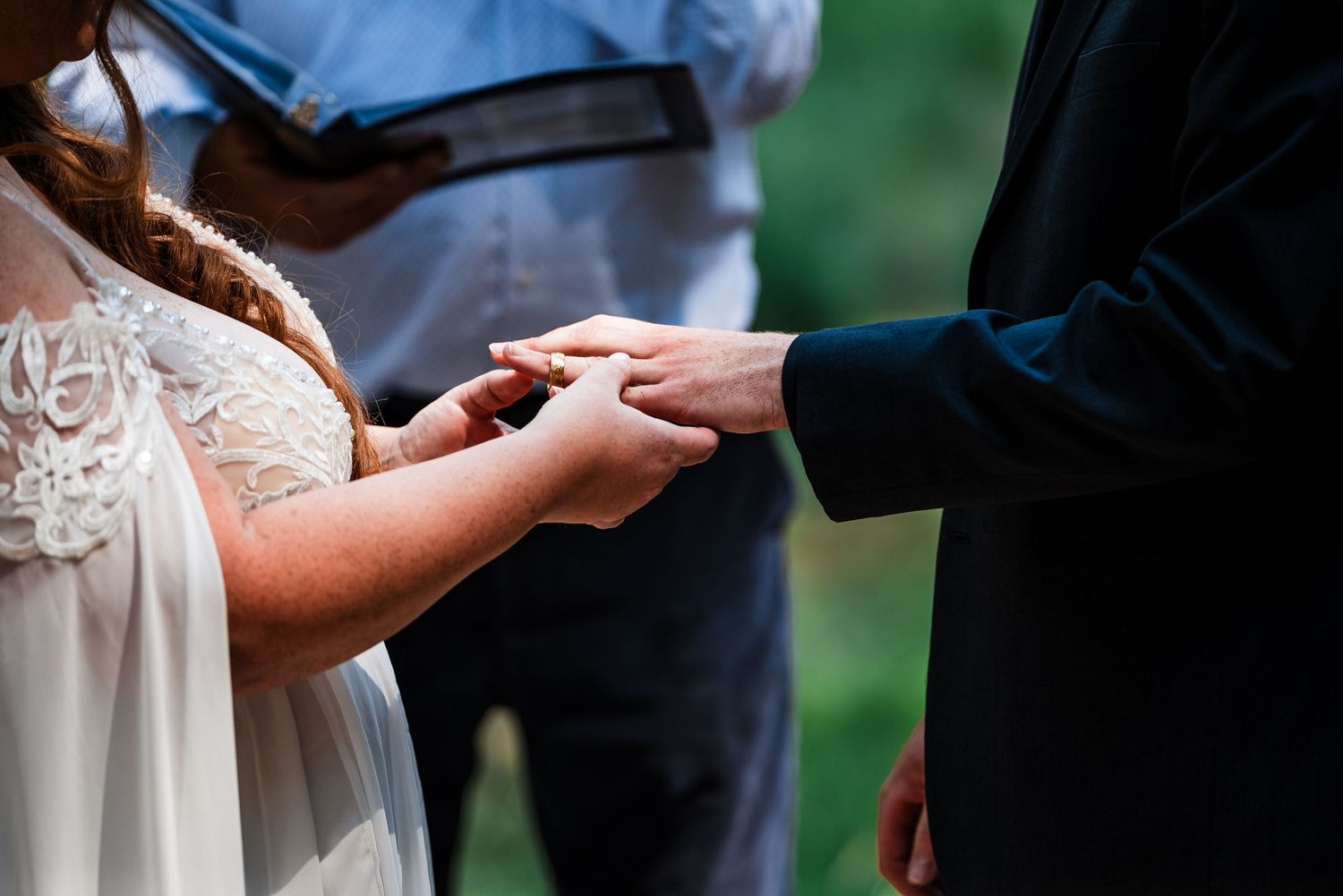 Close-up shot of hands exchanging rings during an outdoor wedding ceremony with greenery in background.